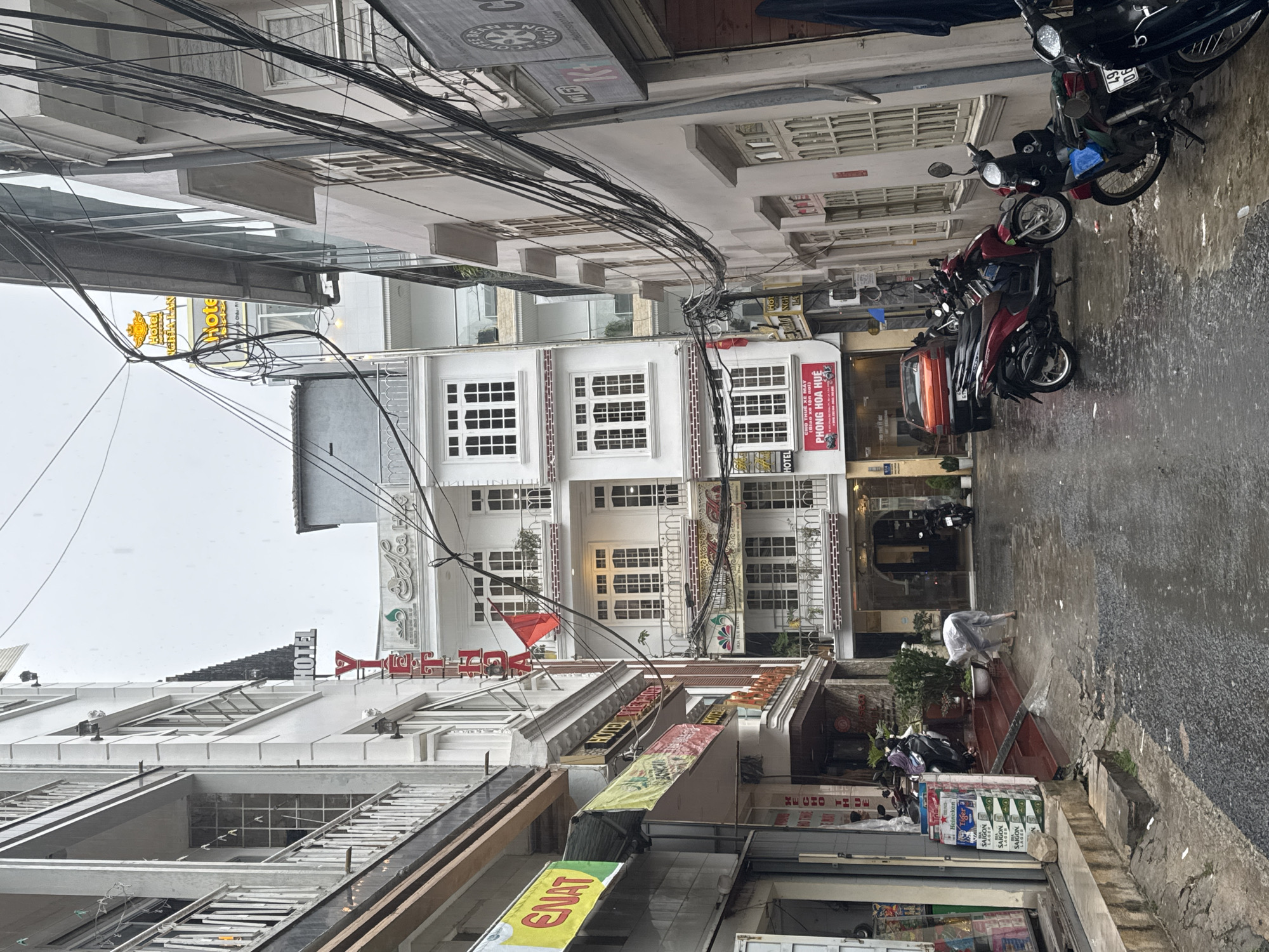 The image depicts a narrow street scene in Da Lat, Vietnam, characterized by a cluster of buildings on either side, overhead cables, and parked motorcycles. The buildings feature a variety of architectural styles and signage, including hotel names and advertisements. A person is seen on the right side of the street, possibly bending over. The street is wet, suggesting recent or ongoing rain.