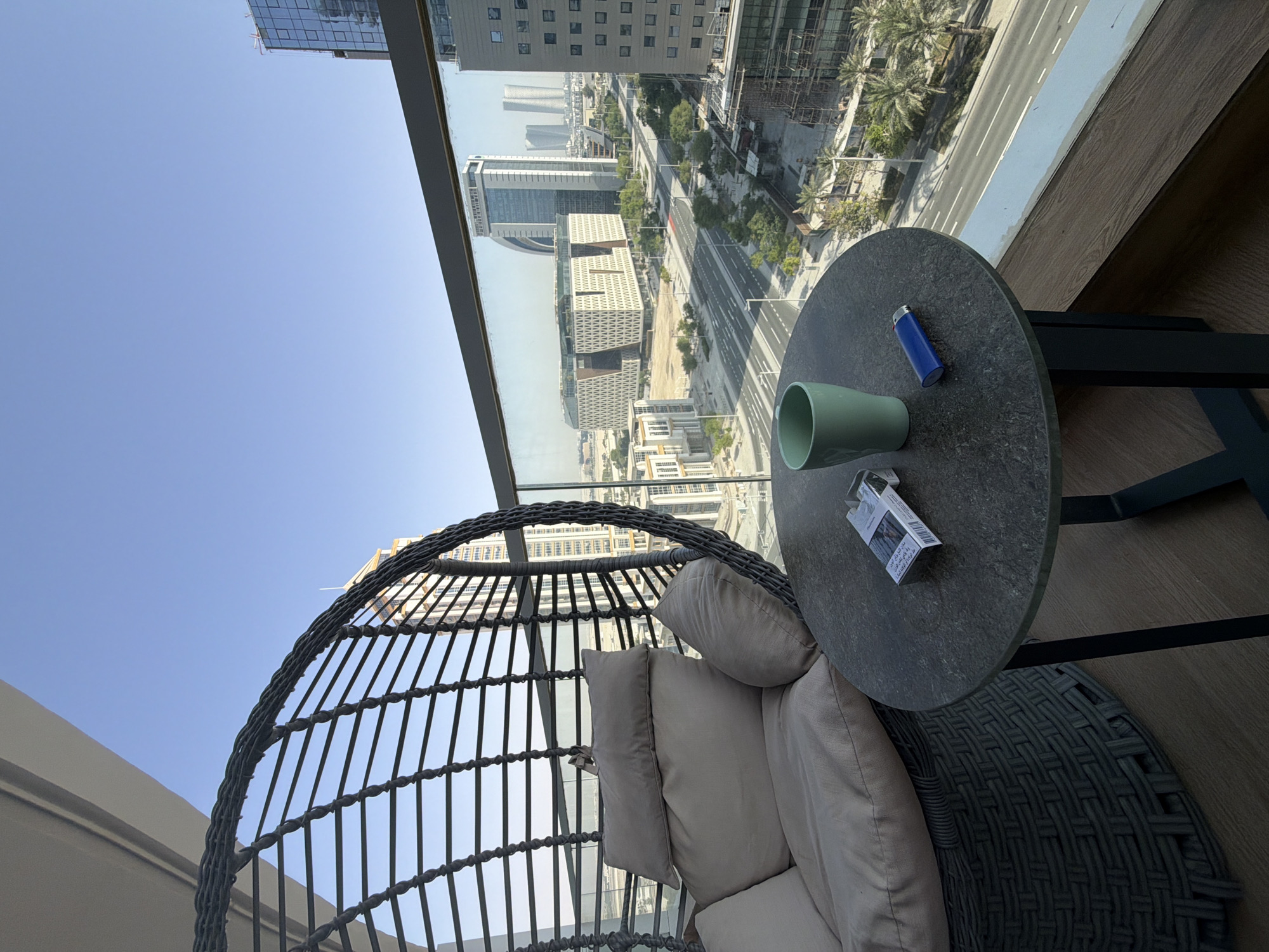 This media file shows an outdoor seating area with a city view. The foreground features a large, round wicker chair with light-colored cushions, appearing cozy and inviting. To the right of the chair, a dark, round table holds a few items: a light green mug, a blue lighter, and a pack of cigarettes with visible text. The table is supported by a dark metal frame.
The background reveals a sprawling cityscape, viewed through a large window or balcony railing. Several modern buildings of varying heights and architectural styles are visible, some under construction. A road with multiple lanes runs through the city, flanked by trees, suggesting an urban environment. The sky is clear and blue, indicating a sunny day, likely morning or afternoon. The overall atmosphere seems peaceful and relaxed, perfect for enjoying the view. The perspective suggests the photo was taken from inside the seating area, looking outwards.