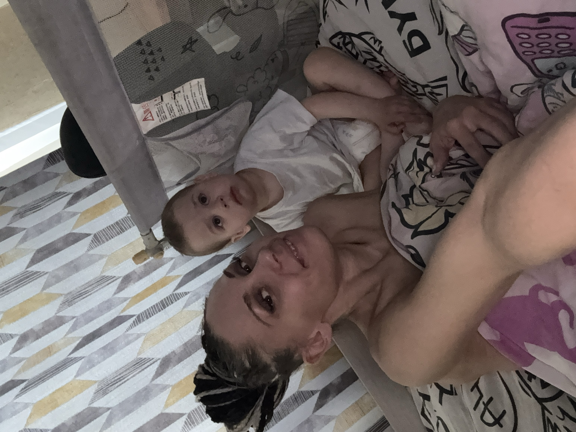 An overhead, tilted indoor shot captures a woman and a young child lying down and looking up towards the camera. The woman, on the right, is partially covered by patterned bedding featuring black text and graphics, including a purple mobile phone. She has dark hair styled in dreadlocks or braids, pulled back, and a slight, relaxed smile. Her bare shoulder is visible.
To her left, a young child, likely a boy, is seen in a white t-shirt. He lies next to or inside a gray mesh playpen or crib, with his head turned towards the camera, displaying a neutral and curious expression. His arm is visible, resting on his shirt.
The floor beneath them features a striking geometric pattern of gray, white, and yellow diamond or chevron shapes. A fabric tag with a red warning triangle and text, including a partially visible "WARNING" and characters in an East Asian language (likely Chinese), is attached to the playpen mesh. A black, rounded object is barely visible under the playpen frame. The lighting is soft, suggesting an indoor setting during the day, creating a calm and intimate atmosphere between the two subjects.