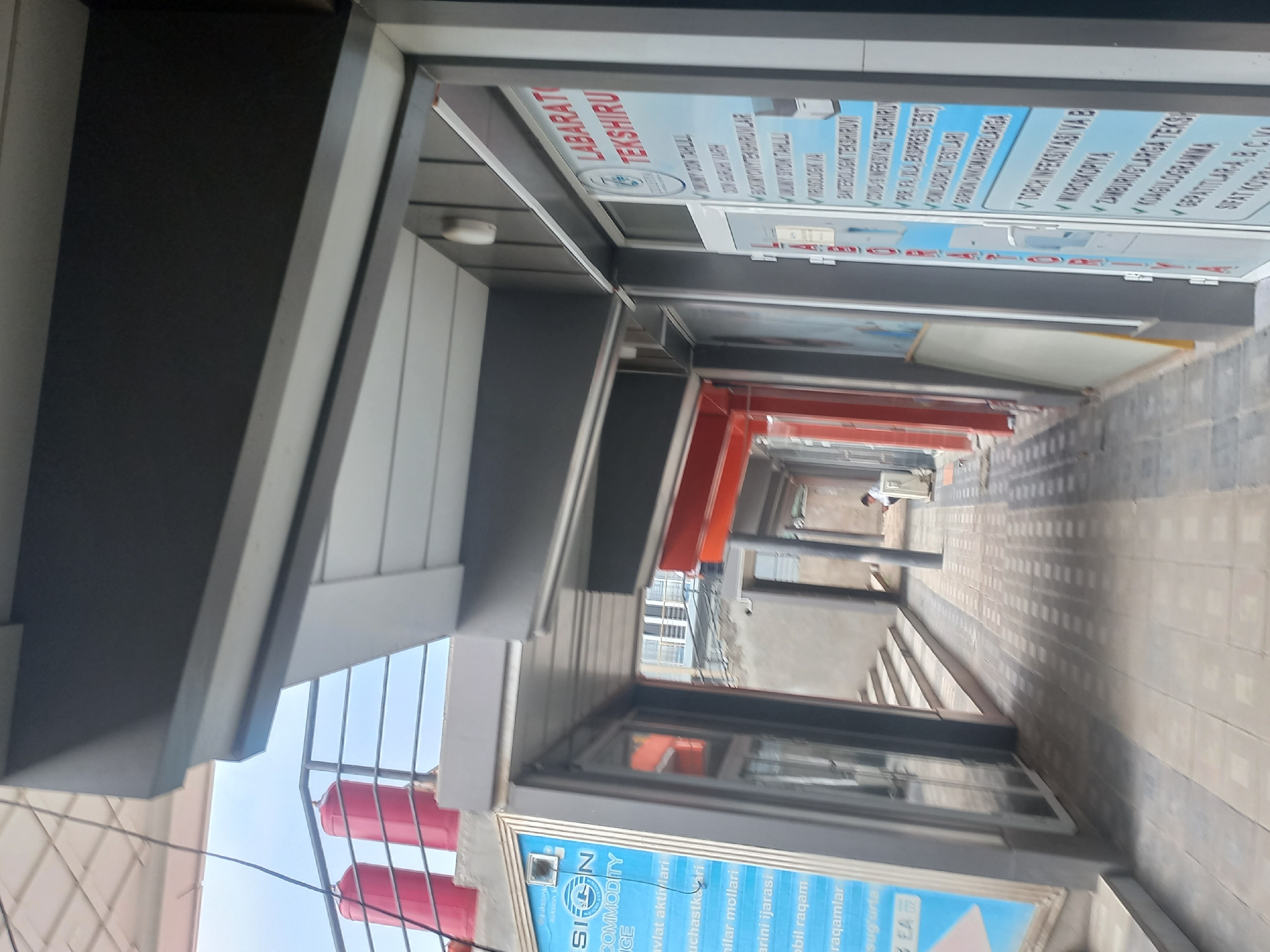 The image shows a street-level view of a building with a covered walkway. Two bright red cylindrical tanks are visible on a raised platform in the upper left of the image, with a metal railing in front of them. To the right of the walkway, there is a storefront with a sign that reads "LABARATORIYA TEXSHIRUV". Below this main heading, there is a list of laboratory tests. The walkway is paved with square tiles, and the scene is lit by daylight. A person is seated on a white box-like object in the background on the right side of the image.