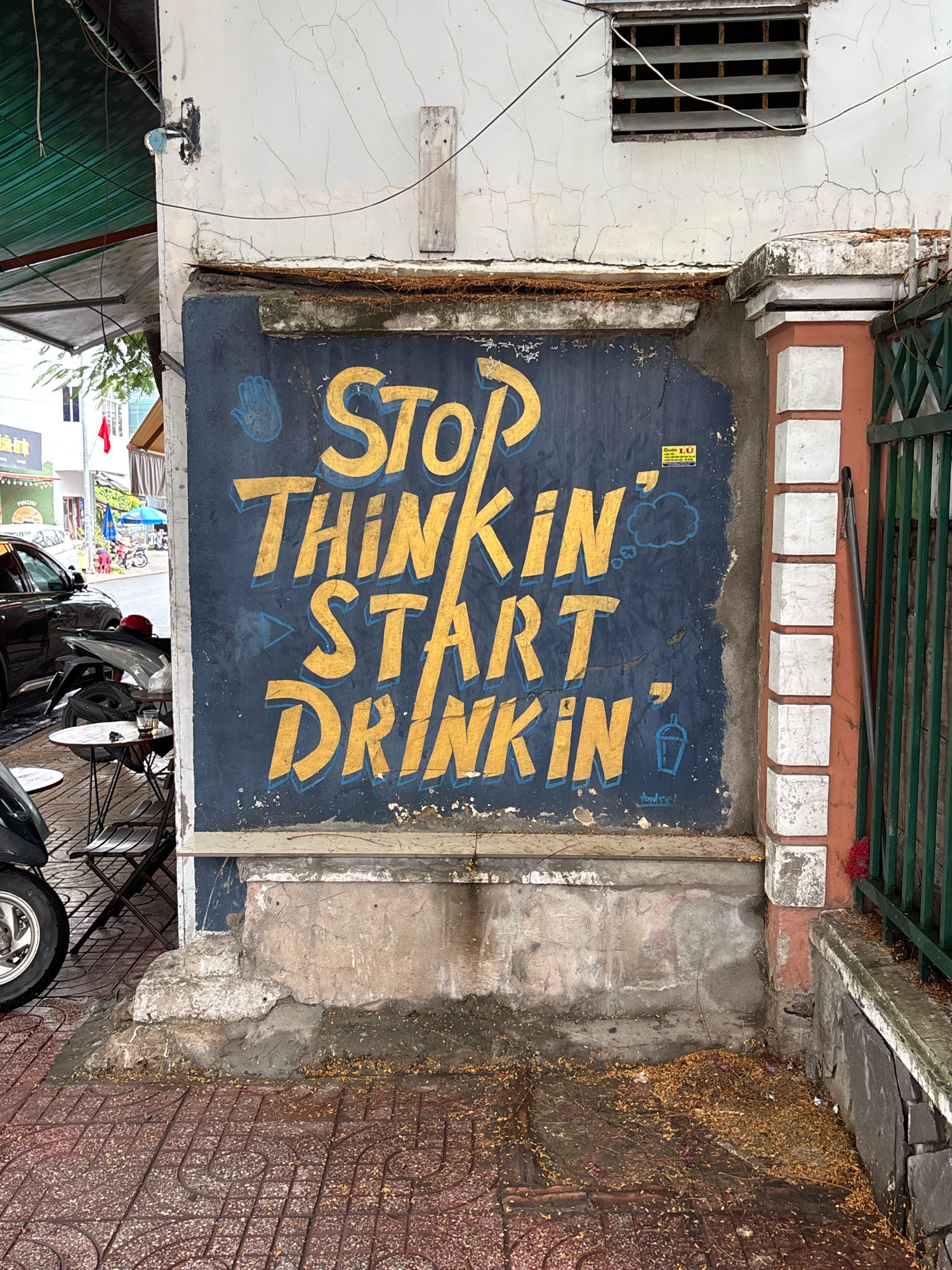 The image shows a weathered blue mural on the side of a building in Nha Trang, Vietnam. The mural features large, yellow, stylized text that reads "STOP THINKIN' START DRINKIN'". There are also blue graphic elements including a hand symbol, a thought bubble, and a cup with a straw. A small yellow sticker with text is visible on the mural. The mural is situated on a dark blue painted wall, which shows signs of age and wear, with cracks and chipped paint. Below the mural, the wall transitions to a rough concrete base. The ground in front of the wall is paved with reddish-brown interlocking bricks, interspersed with debris of fallen leaves and dirt. To the left of the mural, a portion of a dark colored motorcycle and some folding chairs and tables are visible, suggesting an outdoor seating area for a cafe or bar. In the background, a street scene with other buildings, parked vehicles, and some greenery can be observed. The overall scene depicts an urban outdoor setting with a focus on a piece of street art.