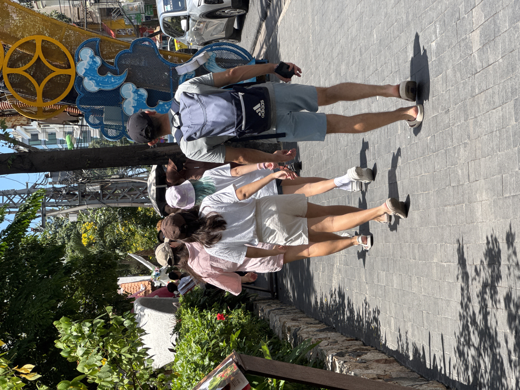 A group of people walk down a paved sidewalk in Nha Trang, Vietnam. The group consists of five individuals, four women and one man, walking in the same direction. The man in the foreground is wearing a grey t-shirt, blue shorts, and a grey backpack with "Adidas" branding. He is holding a black object in his right hand. The women are wearing casual summer clothing, including shorts, t-shirts, and a pink striped shirt. One woman wears a light purple hat, and another a brown baseball cap. They are walking past lush green foliage and stone-lined planters on the left side of the sidewalk. In the background, buildings and electrical wires are visible, along with a brightly colored decorative structure featuring a yellow circular emblem and blue wave motifs. A white vehicle, possibly a taxi, is parked on the right side of the street. The ground is paved with dark grey interlocking bricks, and shadows from trees and overhead structures are cast across the path. The sky is bright and appears to be clear.