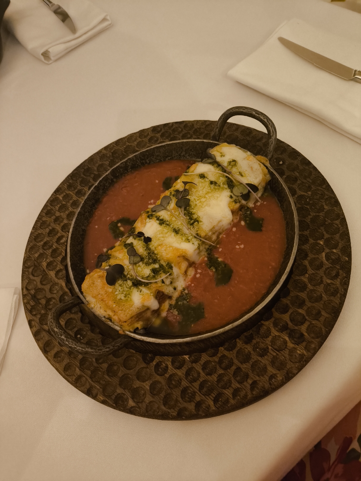 A high-angle shot captures a dish of food served in a rustic pan on a wooden trivet, presumably at a restaurant. The dish appears to be layered with melted cheese, a green herb garnish, and served in a bright red tomato sauce. The trivet has a pattern of raised dots and is placed on a white tablecloth. To the upper right and left, white napkins are visible, with a single knife resting on one. The location is Unknown, Unknown.