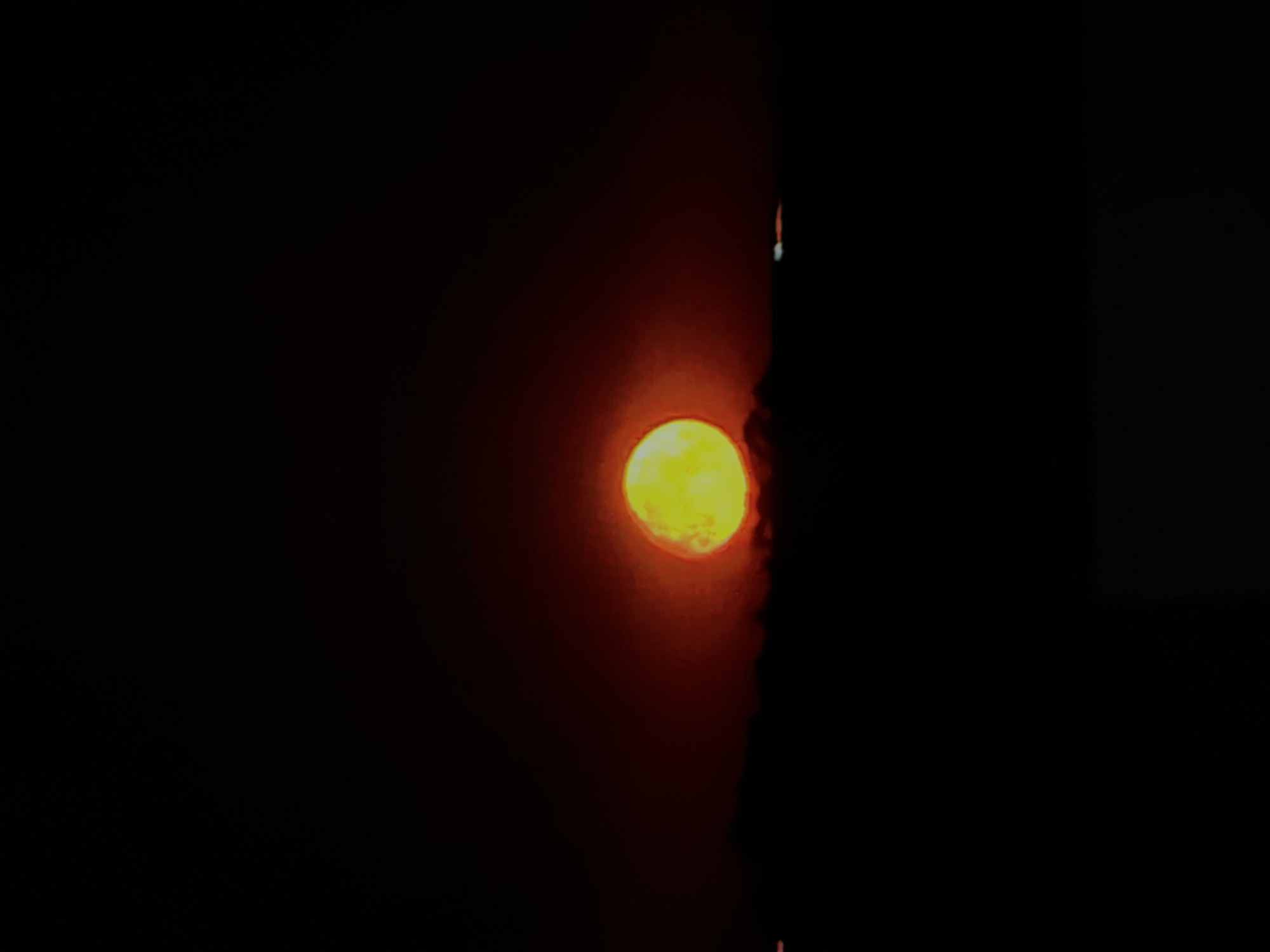 A bright yellow-orange sun is low in the sky, partially obscured by a dark, silhouetted horizon. The sky is a deep, dark hue, with hints of red and orange around the sun, suggesting it is either sunrise or sunset. The horizon is flat and indistinct, indicating a vast open area. There are a few faint lights visible on the horizon, possibly from distant buildings or vehicles. The overall mood is serene and dramatic.