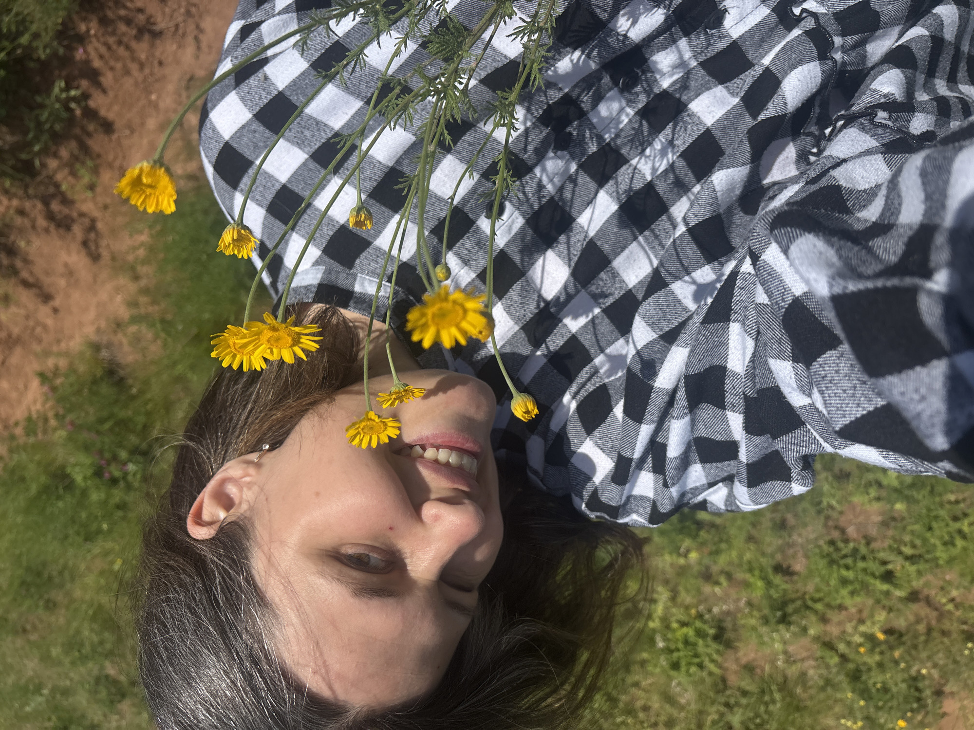 A young woman with dark hair and fair skin is shown in a close-up, angled portrait. She is wearing a black and white checkered shirt and is smiling, with a slight wink. She is holding a small bunch of yellow wildflowers. The background is a blurred expanse of green grass and brown earth, suggesting an outdoor, natural setting. The lighting is bright, indicating it is daytime.