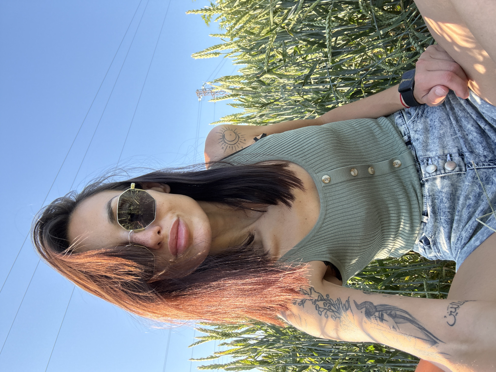 A woman is sitting in a field of tall grass under a blue sky. She is wearing a light green ribbed tank top, denim shorts, and sunglasses. She has dark hair with auburn highlights. The woman has tattoos on her arms and shoulder. There are power lines visible in the background. The location is Pleven, Bulgaria.