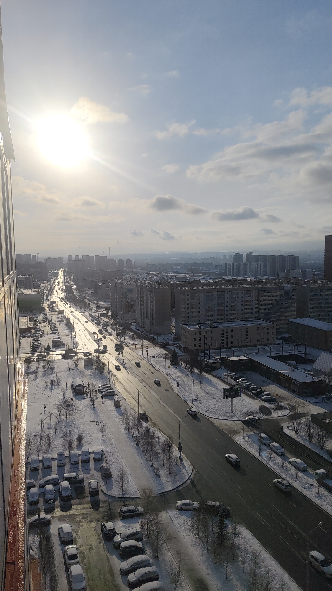 A high-angle, daytime view of a winter scene in Krasnoyarsk, Russia. A major city street runs through the center, with vehicular traffic visible. The street is mostly clear of snow, but adjacent sidewalks and parking areas are snow-covered. Many cars are parked along the street and in designated parking lots. Buildings of varying heights are present, mainly residential apartment blocks. Trees are bare, indicating the season. People are visible as small figures in the snow-covered areas. The sky is partly cloudy, with the sun appearing bright. A building edge is seen in the left foreground, indicating the viewpoint is from an elevated position inside a building.
