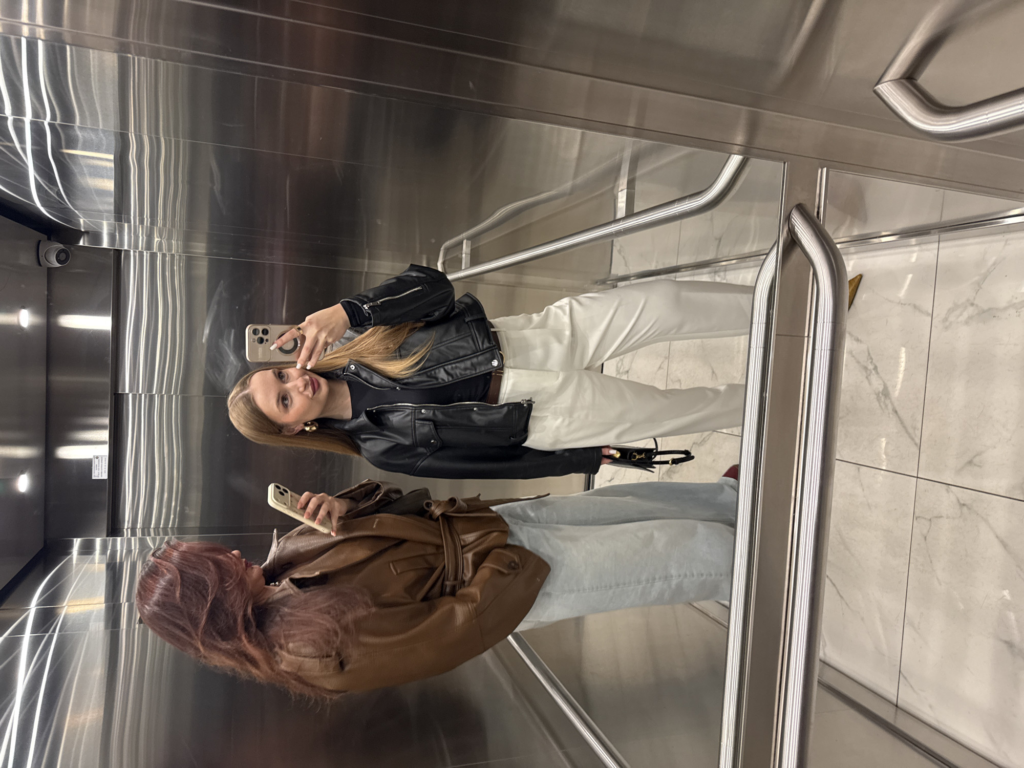 Two women stand in an elevator, as reflected in the mirrored wall. The woman at the top of the frame is blonde and wears a black leather jacket, white pants, and black shoes. She holds a phone in her right hand and takes a selfie. The woman in the lower part of the frame has red hair, wears a brown leather jacket and blue jeans. She holds a phone in her right hand and takes a selfie. The elevator's interior is primarily stainless steel with metal handrails. The floor is patterned with what appears to be white marble tiles. A security camera is visible in the upper left of the frame. The scene appears to be inside an elevator. Due to the lack of visible landmarks, it's impossible to confirm the exact location within the city of Belgrade, Serbia.