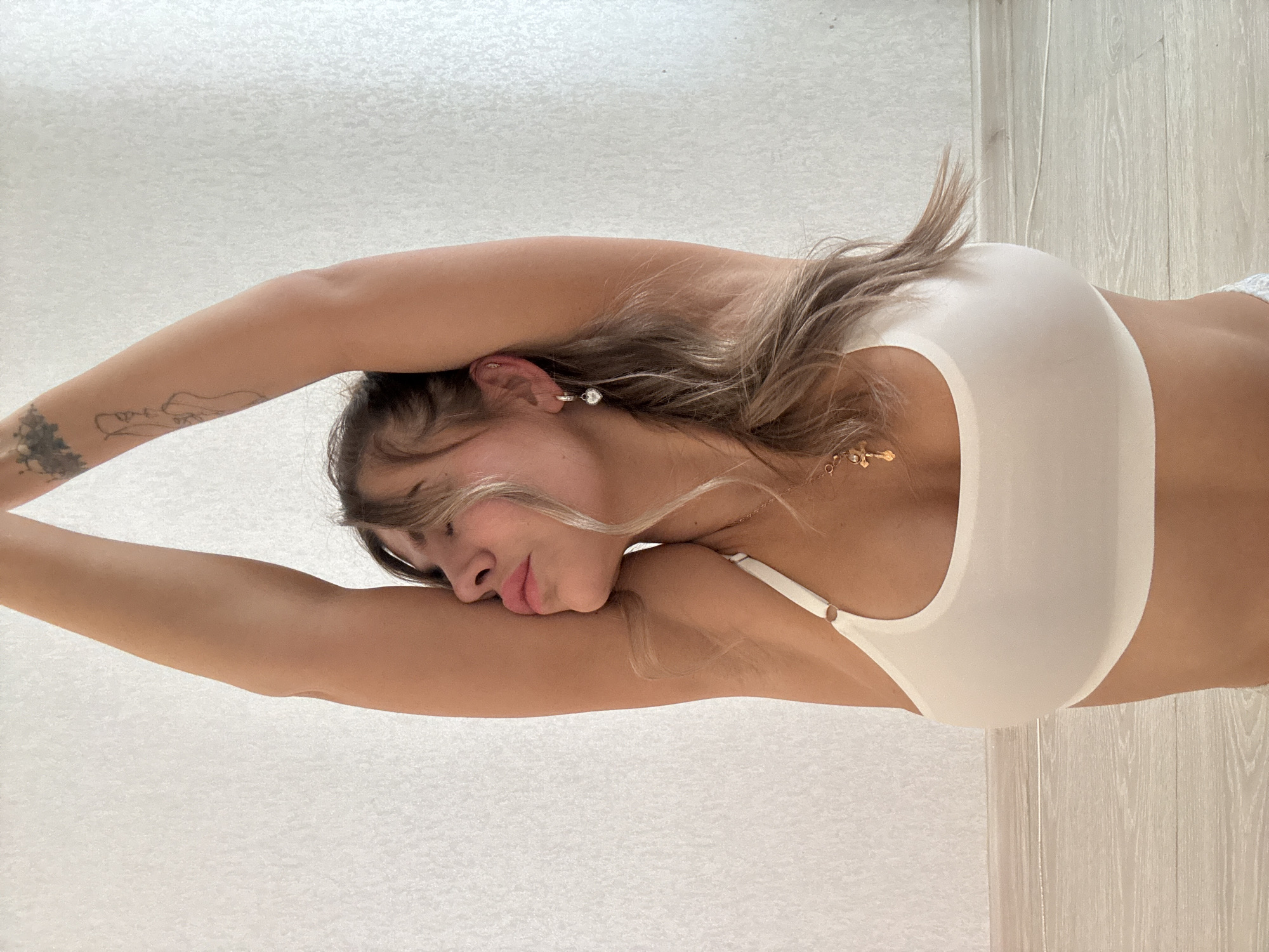 The image shows a female subject positioned indoors, engaged in a static pose consistent with stretching or yoga. The subject, exhibiting a medium skin tone, has her eyes closed, head tilted slightly upwards and to her right, with her mouth slightly open. Her lips appear full and tinted pink. Both arms are raised vertically overhead, bent at the elbows. She wears a plain white sports bra. A gold-colored necklace with a cross pendant is visible. In her right ear, a silver-colored hoop earring with a small heart-shaped charm is present, along with an upper cartilage piercing. Tattoos are visible on both upper arms: a faded dark design on the left, and a fine-line graphic on the right. The background is a plain, light-colored textured wall, and a light-toned wooden floor is visible at the bottom of the frame. No specific visual identifiers are present in the image to confirm a location within the city of Chaikovsky, Russia.