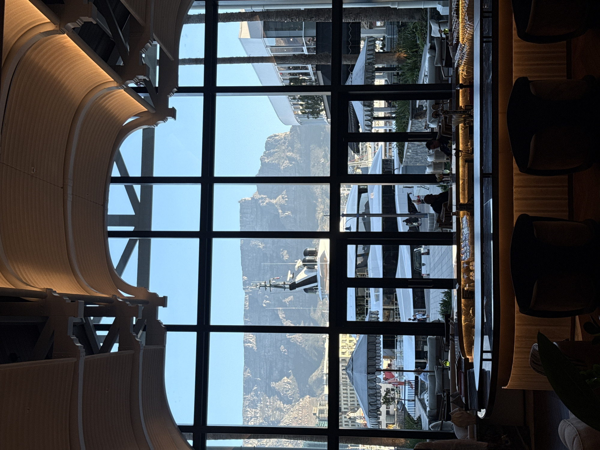 The image shows an interior view through a large window with a grid pattern. Outside, Table Mountain dominates the skyline. In the foreground, a bar area is visible with chairs and tables, suggesting a restaurant or lounge setting. People are seated at the bar. Through the window, a marina is visible with several yachts. The scene depicts Cape Town, South Africa, with Table Mountain as a prominent feature. The image includes architectural elements of the interior, with a light-colored, textured ceiling. The light suggests it is daytime.