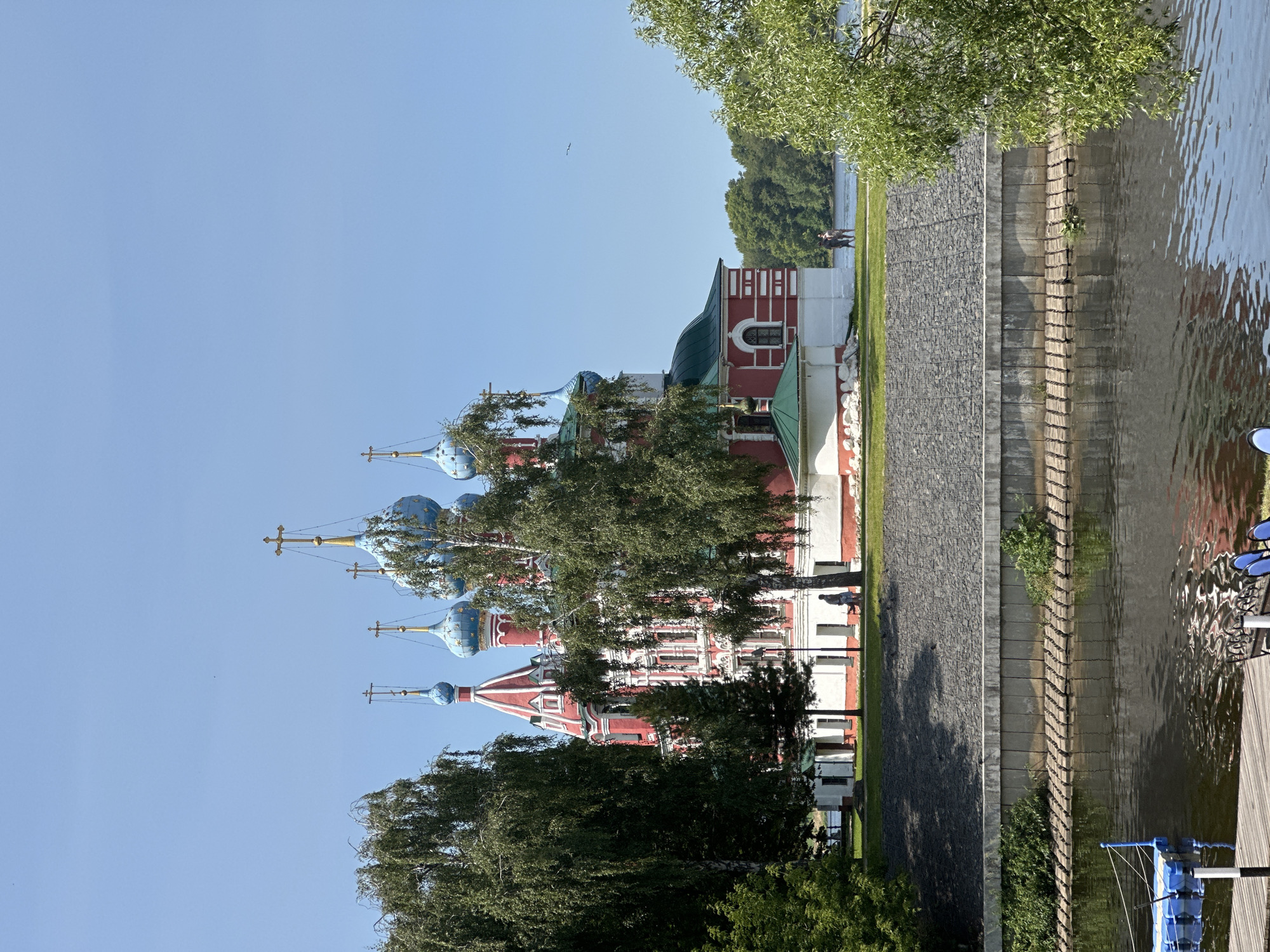 The image shows a view of the Church of the Smolensk Icon of the Mother of God in Terpilovo, Russia. The church, a prominent architectural landmark, is situated on a hill overlooking a body of water. It features a vibrant red and white facade with numerous blue domes topped with golden crosses. Lush green trees partially obscure the church, framing the scene.
In the foreground, a wooden dock with blue seating and a blue floating platform is visible. The water reflects the sky and surrounding foliage. A stone embankment separates the water from the grassy area where the church stands. A few individuals are seen walking on the grass near the church. The sky is clear and blue, indicating a sunny day. The overall impression is one of a serene, picturesque landscape dominated by the distinctive religious architecture.