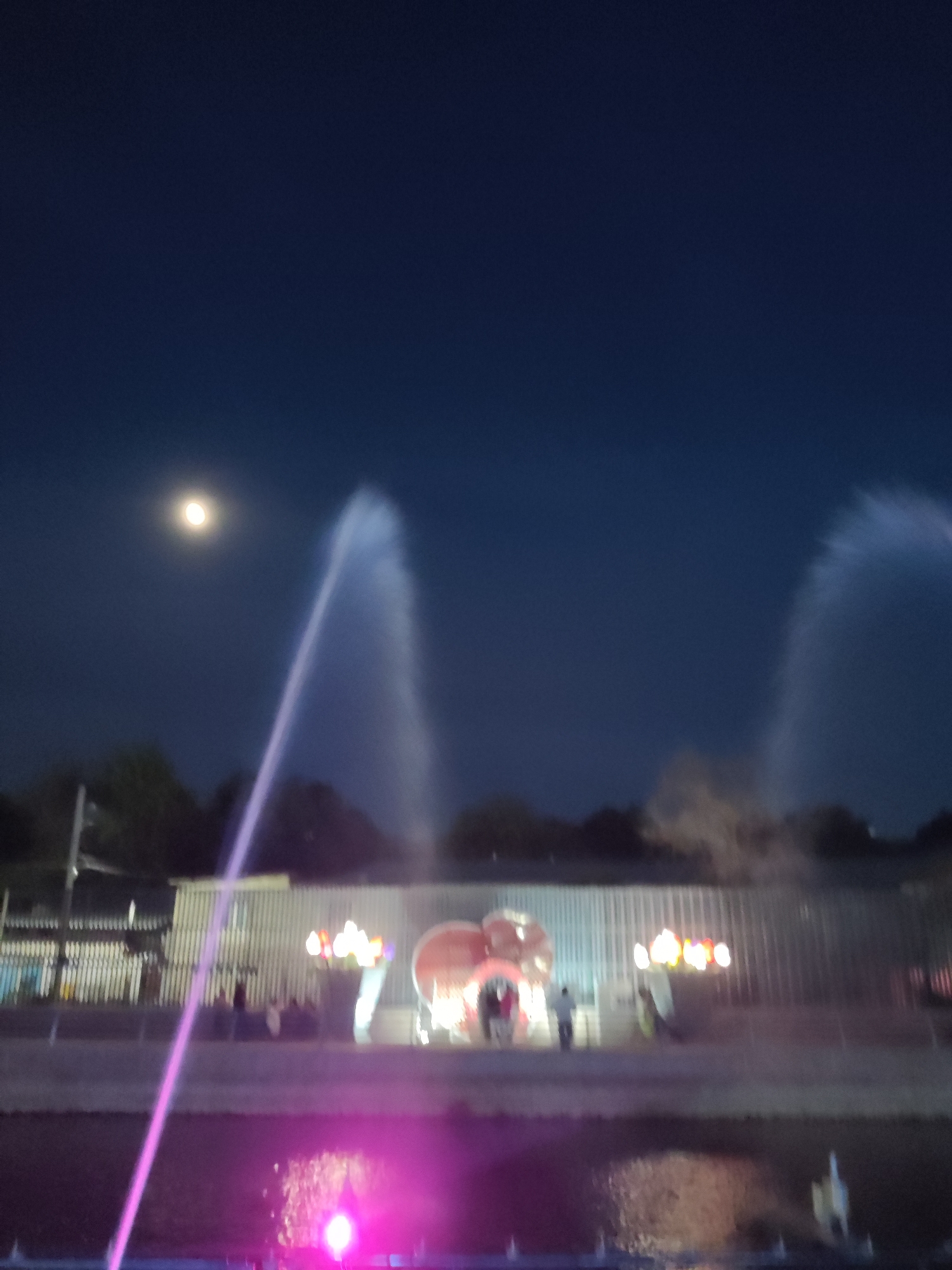 An outdoor nighttime scene features a large, illuminated fountain and a building structure. The sky is dark blue, with a bright, full moon visible in the upper left quadrant, to the left of a tall, arcing water spray from the fountain. Several other water sprays are visible, some appearing as plumes of mist.
In the foreground, the water of the fountain is illuminated with vibrant pink and purple lights, reflecting on the surface and creating bright spots. A long, thin beam of pink light, likely from a laser or light projector, extends diagonally from the lower left corner towards the center of the image.
Behind the fountain, a long, horizontally oriented building is visible. It has a corrugated metal exterior and is illuminated by various colored lights, including strings of yellow and red lights above a decorative structure that appears to be heart-shaped. Several blurry figures of people are present in front of the building, some appearing to be interacting with the lights or structures. The overall atmosphere suggests a public space, possibly a park or entertainment venue, at night. The location is identified as Shymkent, Kazakhstan.