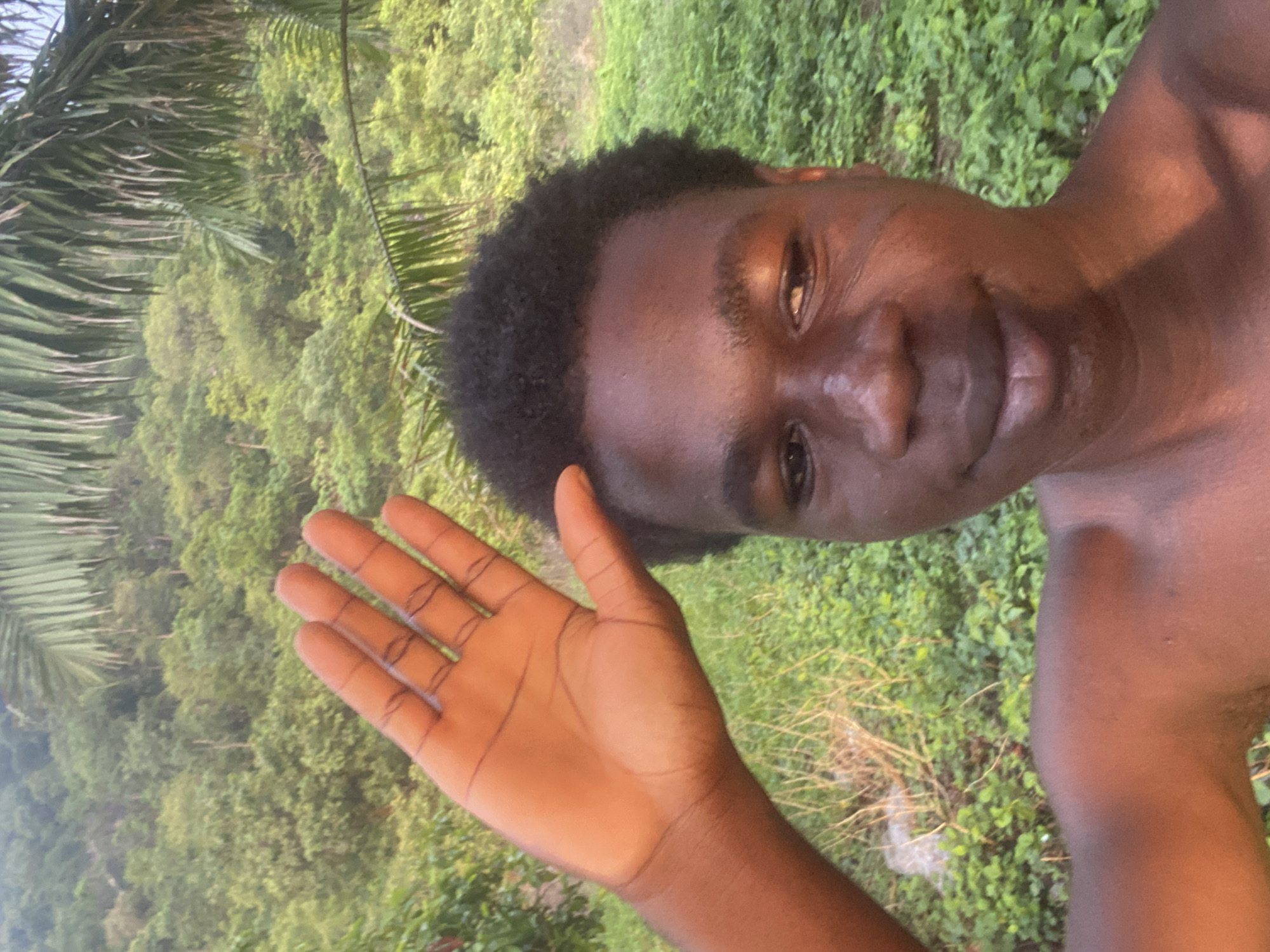An individual with dark skin and short, dark hair is posing for a selfie in an outdoor setting. The person is shirtless, with a hand raised in a waving gesture. The background appears to be a lush, green area with trees and foliage. The specific location in Mararaba, Nigeria, is not discernible from the image alone.