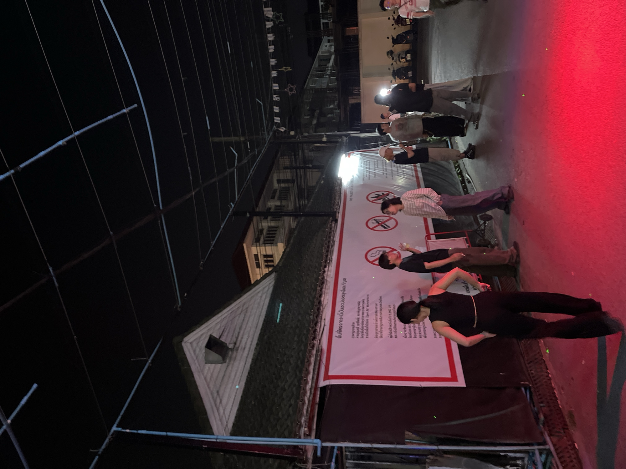 The image shows a night scene in Chiang Mai City Municipality, Thailand. People are standing or walking on a red sidewalk next to a white sign with Thai writing and red circles with crossed-out cigarettes on it, signifying a no-smoking area. Buildings and overhead wires are visible in the background. There are also scooter parked in the background. The lighting is dim, with some artificial lights illuminating the scene.