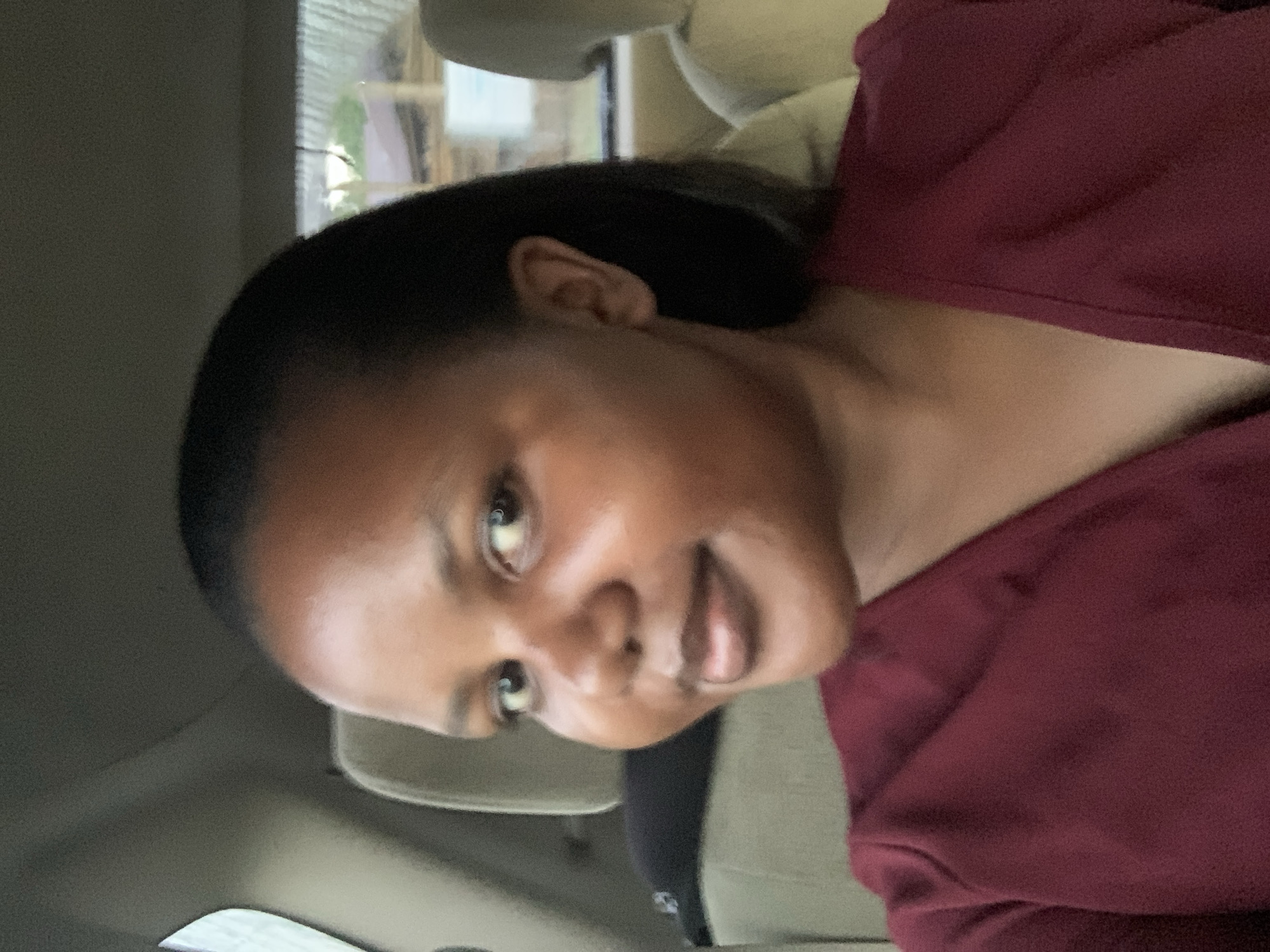 A medium shot depicts a dark-haired woman in a maroon shirt, seated in a vehicle. The setting is indicated as Dar es-Salaam, Tanzania, but specific environmental details are limited to the interior of the car and a glimpse of the exterior through the window. The woman faces the camera with a slight smile. No specific actions or interactions are discernible.