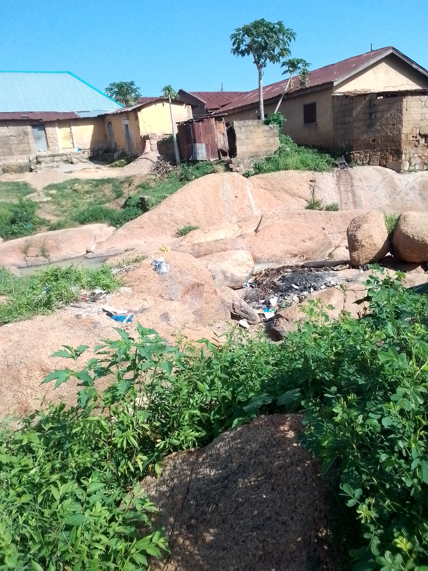 This image depicts a rural or semi-rural environment in Jos, Nigeria. In the background, several buildings with corrugated iron roofs and mud brick walls are visible. Some of these structures appear to be residences or small shops. The foreground is dominated by large, rounded rocks, possibly part of a natural rock formation. Lush green vegetation, including bushes and trees, grows around and on these rocks, with some areas appearing overgrown. In a low-lying area between some of the rocks, there is evidence of refuse and debris, including what appears to be burnt material and scattered trash. A small amount of water is visible in a channel or depression amongst the rocks. The sky is clear and blue. The scene suggests an area that is not well-maintained, with a mix of natural landscape and human habitation.