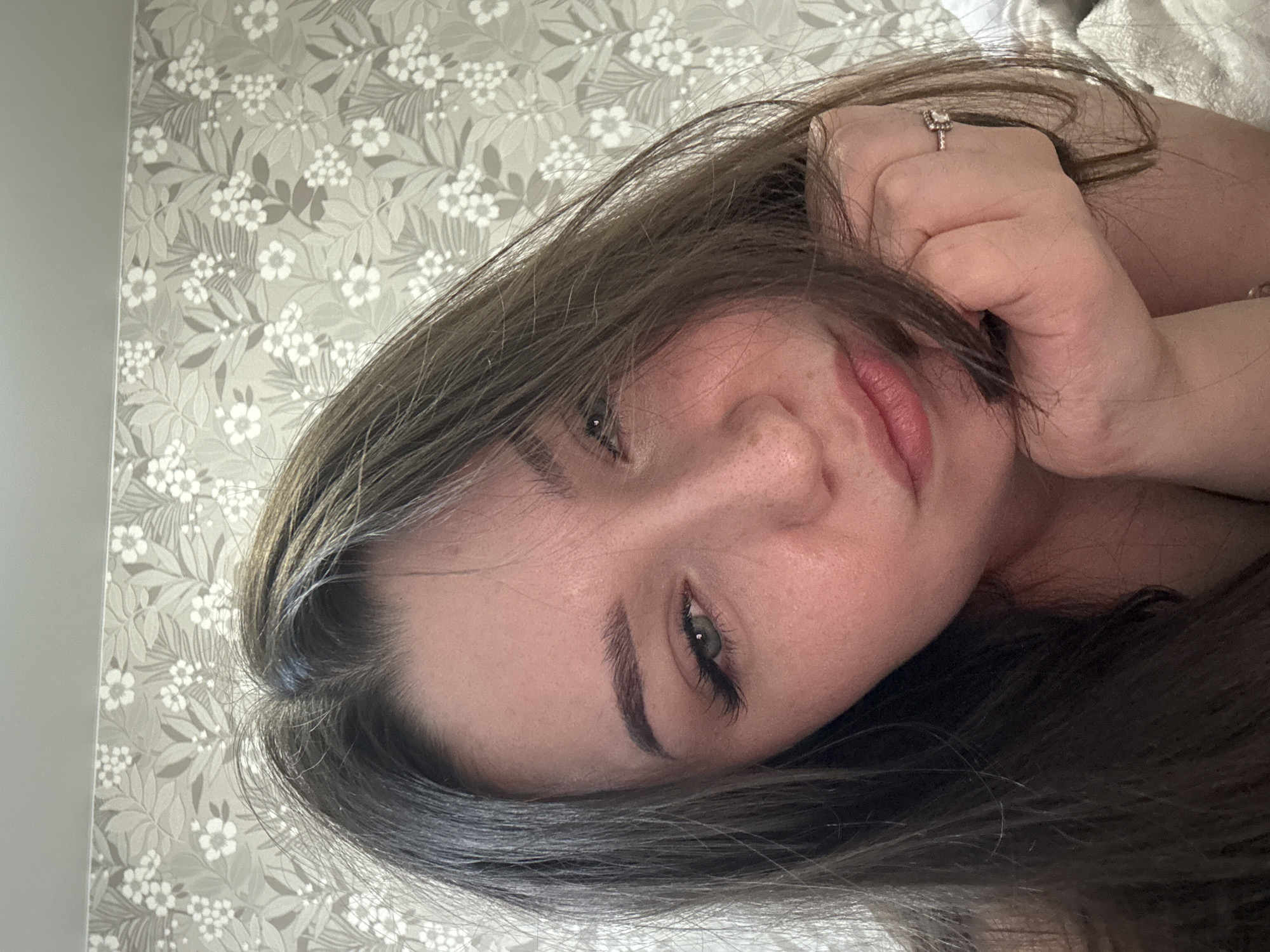 The media file is a close-up, over-the-shoulder selfie of a young woman lying down, with her head tilted and her hand supporting her chin. She has long, dark brown hair that falls around her face and shoulders. Her eyes are a striking green and appear to be looking directly at the camera. She has on light makeup, including mascara and possibly some subtle eyeshadow. Her lips are slightly parted, revealing a soft pinkish hue. A ring with a rectangular stone is visible on her right hand. The background features a wallpaper with a light gray base and a repeating pattern of small white flowers and green leaves, suggesting an indoor setting, possibly a bedroom given the soft lighting and the texture of what appears to be a duvet or blanket behind her. The lighting suggests it could be daytime, with natural light filtering into the room. The woman's expression is calm and relaxed, bordering on contemplative. The overall mood is intimate and personal.