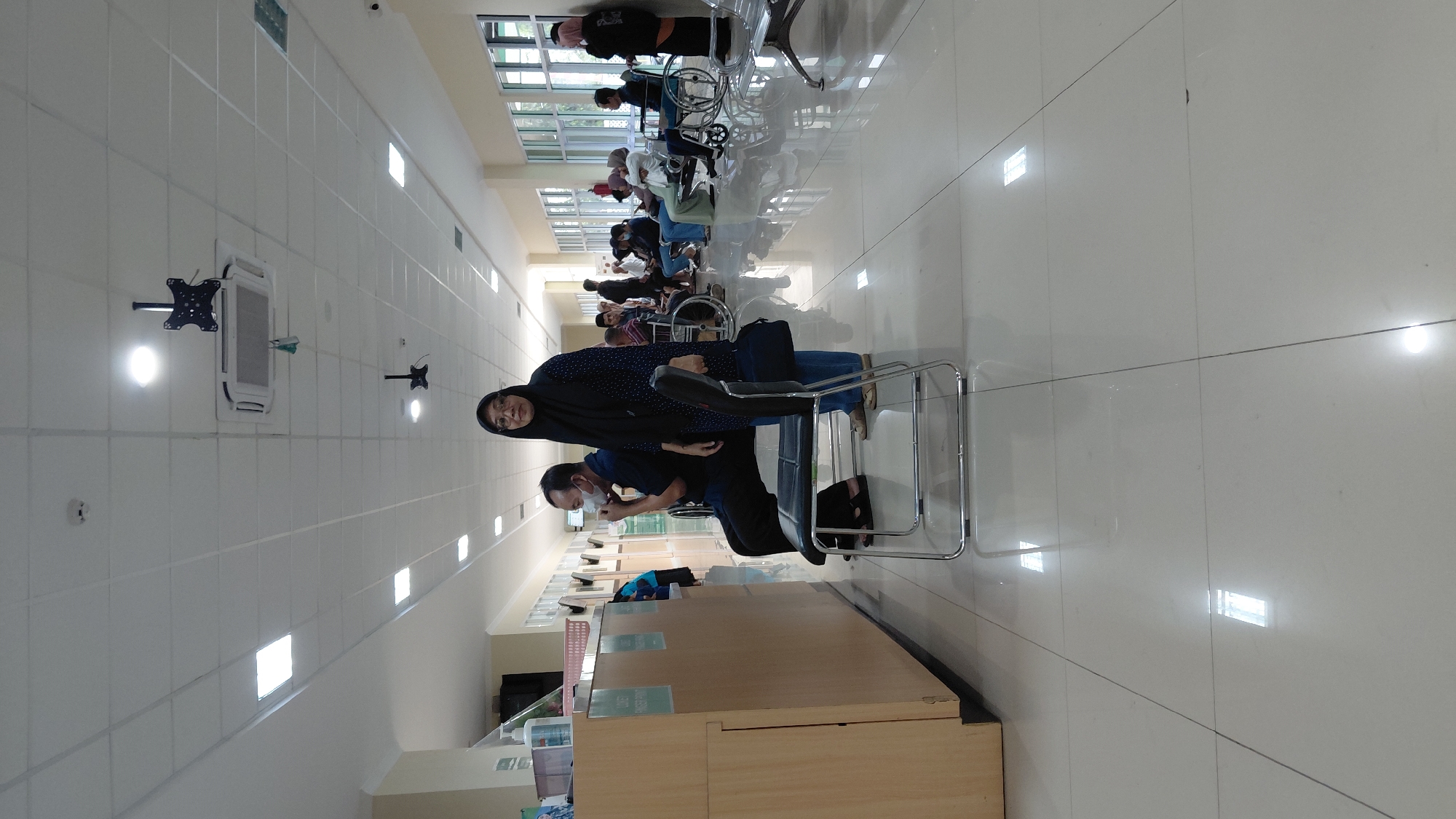 The image depicts a brightly lit, modern waiting area, likely within a healthcare facility in Rejasari, Indonesia. The floor is tiled with large, reflective white tiles. The walls are a pale cream color, and the ceiling is a suspended white tile system with numerous embedded fluorescent lights.
In the foreground, a man wearing a blue shirt and dark pants sits on a metal chair with a black seat, his face partially obscured by a white mask. To his right, a woman wearing a dark hijab and a patterned dark blue and white top stands, holding a dark bag. She is looking towards the camera. Behind them, a wooden counter with various items, including what appears to be hand sanitizer, is visible.
Further back in the waiting area, several other individuals are seated or in wheelchairs, suggesting a patient waiting room. Many are dressed in casual or modest attire, consistent with local customs. Windows line the far wall, letting in natural light. In the distance, another person in a dark hijab stands near a window. The overall atmosphere is calm and orderly.