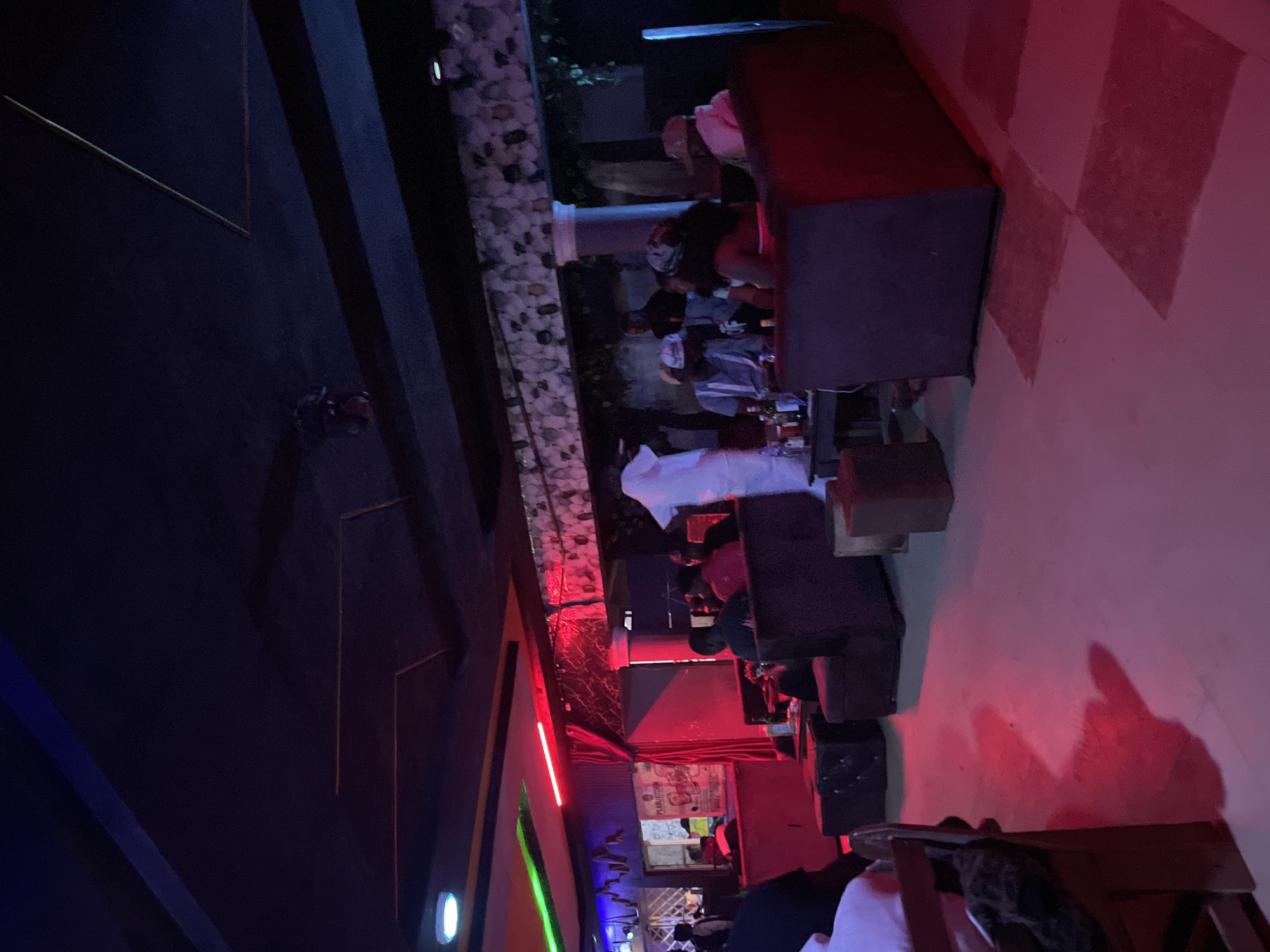 The image depicts an indoor scene at night, illuminated by red and green neon lights and ambient lighting. The setting appears to be a lounge or club in Durumi, Nigeria. Several individuals are present, seated on plush sofas and chairs arranged in a lounge area. Some are engaged in conversation, while others are observing their surroundings. A table in the foreground holds several bottles, suggesting a social gathering. The decor includes textured walls and a wooden chair. The overall atmosphere is dimly lit and colorful, typical of a nightlife venue.