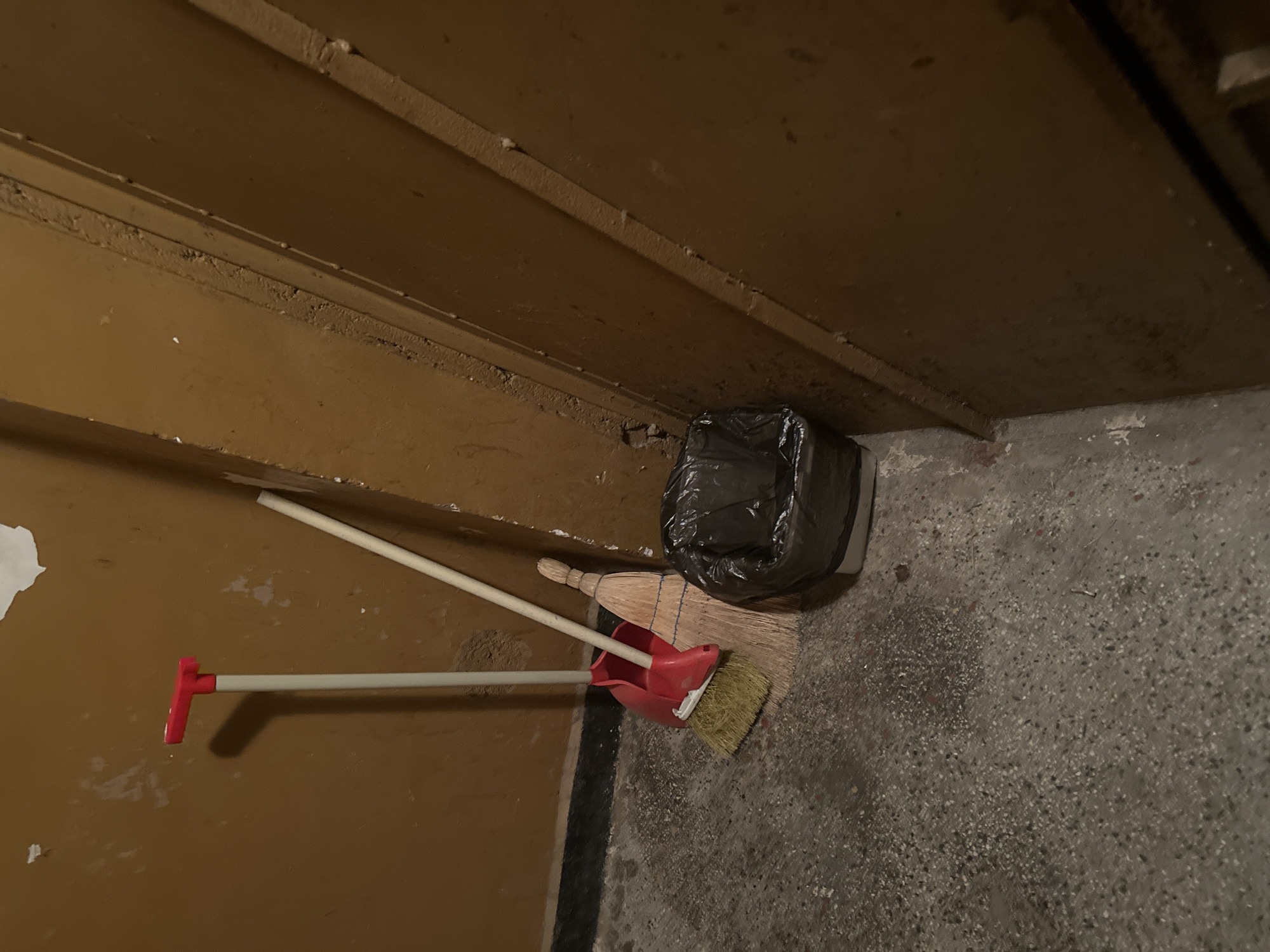 This image captures a close-up view of a corner in what appears to be a functional, possibly neglected, indoor space in Blagoevgrad, Bulgaria.
The scene is dominated by drab, ochre-brown walls, which could be painted wood panels or plastered surfaces, showing signs of wear and some peeling paint or plaster debris, particularly on the left side. The floor is made of rough, speckled concrete with various shades of grey and dark aggregate visible.
In the corner, two brooms and a dustpan are stored. One is a traditional straw broom with a light-colored handle. Next to it is a smaller broom, also with a light handle, paired with a bright red dustpan. These cleaning tools lean against the wall and are somewhat intertwined. Further into the corner, a black plastic garbage bag, appearing full and crumpled, rests on the concrete floor against the wall.
The lighting is dim, suggesting an interior location such as a utility room, a basement, or a service area. There is no visible human activity or specific event taking place; the items are static and stored. No people are present. The atmosphere is utilitarian and somewhat unkempt. No text is visible anywhere in the image.