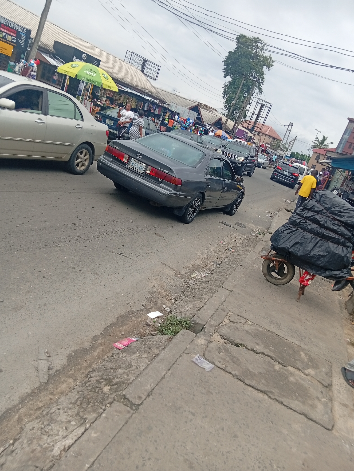 The image depicts a street scene in what appears to be a busy market area. The foreground shows a gray Toyota Camry car in traffic. A person in a yellow shirt is standing near a red wheelbarrow loaded with black plastic-wrapped items on the right side of the road. In the background, there are several other cars, motorcycles, and a market with stalls and umbrellas. The sky is overcast. The road is paved and shows signs of wear. There is also some debris on the side of the road.