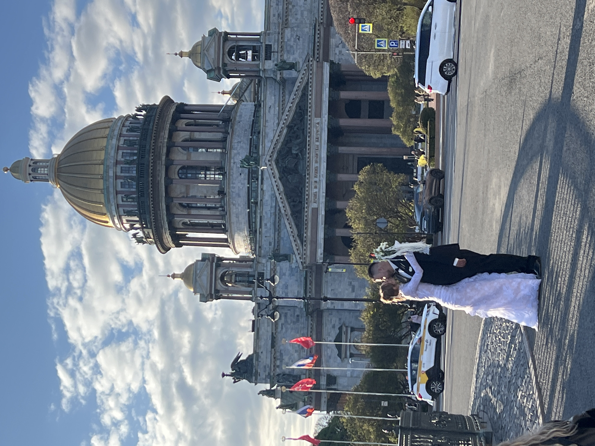 The image depicts a wedding scene in Saint Petersburg, Russia, in front of Saint Isaac's Cathedral. The bride and groom are in the foreground; the groom is wearing a black suit, and the bride is in a white wedding dress. They appear to be kissing. Several flags are on display, and white vehicles are parked nearby. The scene is set against a partly cloudy sky.