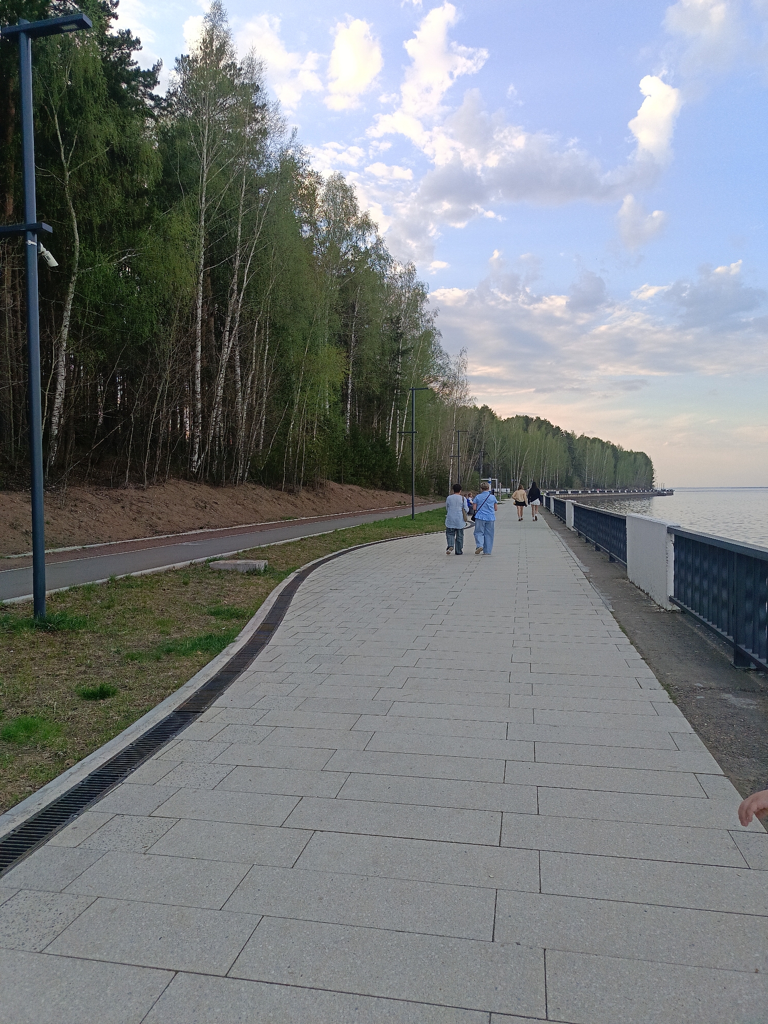 The scene is a pedestrian walkway in Chaikovsky, Russia, alongside a body of water, likely the Kama River. A group of people are walking along the paved path. The walkway is constructed from large rectangular stones and includes a drainage grate along the edge bordering a grassy area and a paved bike path. The background consists of a dense forest line, and a metal railing separates the walkway from the water. The sky above is lightly cloudy. A hand is visible in the lower right corner of the picture.