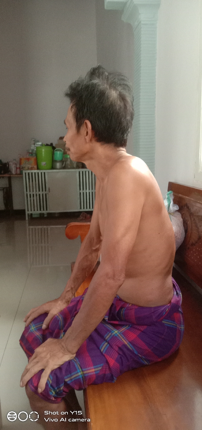 The scene depicts an interior residential space located in Brumbungan Kidul, Indonesia. A bare-chested adult male, appearing middle-aged or older, is seated in profile, facing left. He wears a purple, red, and blue plaid sarong. His hands rest on his knees. He occupies a light brown wooden bench or chair, positioned against a light-colored wall. In the background to the left, a white metal cabinet with slatted doors is visible, topped with a green lidded plastic container and other miscellaneous objects. To the right, a window with a white frame admits natural light, with sunlight evident on the man's right shoulder. A white plastic bag containing indistinct items is positioned on the wooden furniture near the window. A light blue, vertically fluted architectural element is present on the far right wall. The flooring appears to be tiled.