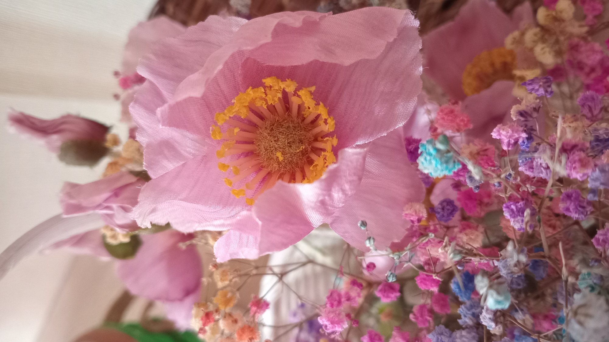 The image is a close-up, slightly angled, vertical shot of a floral arrangement. The dominant subject is a large, pink, fabric flower with delicate, slightly frayed petals. Its center features a cluster of yellow stamens and a brownish-tan pistil. To the left and below this large flower, a bed of smaller, multi-colored dried baby's breath flowers is visible. These smaller flowers are in shades of pink, purple, light blue, and beige, with thin, dry stems. The background is out of focus, showing hints of a textured surface, possibly a basket weave and a light-colored fabric, suggesting the arrangement might be in a container or part of a larger decorative piece. There is no indication of a specific location within Slobozia, Moldova, or any discernable human presence or action. The scene appears to be a still-life arrangement of artificial or dried flowers.