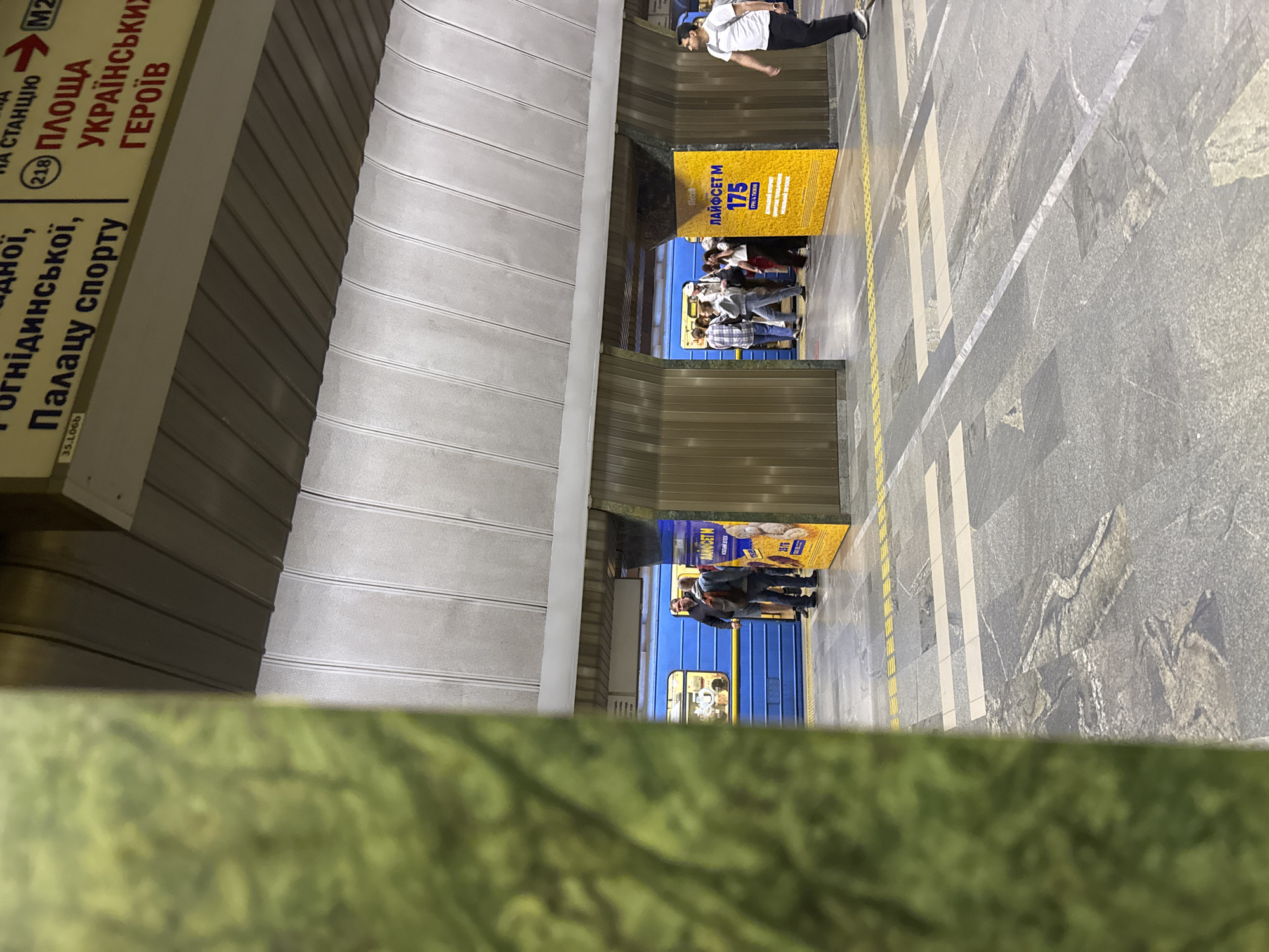 The image captures a scene within a metro station in Kyiv, Ukraine, presented from a rotated perspective (90 degrees clockwise).
The scene shows a subway platform with a blue train stopped at the station. The platform features a patterned grey tiled floor with distinct white and yellow line markings, including a dashed yellow line along the edge. The station's architecture includes paneled walls and a textured, possibly ribbed, ceiling.
Several people are visible on the platform. One individual, dressed in a white t-shirt and dark pants, stands further down the platform. A group of people is gathered near the open doors of the blue subway train, appearing to be in the process of boarding or disembarking. Another small group is seen further along the platform. The overall activity suggests routine transit within the metro system.
Notable details include overhead signage in Ukrainian, which points "НА СТАНЦІЮ М2" (TO STATION M2) and lists destinations such as "(218) ПЛОЩА УКРАЇНСЬКИХ ГЕРОЇВ" (Ploshcha Ukrayins'kykh Heroiv - Ukrainian Heroes Square) and "Палацу спорту" (Palace of Sports). This clearly identifies the location as a metro station in Kyiv. Two large yellow advertising boards are prominent on the platform, displaying the text "ЛАЙФСЕТ М" (LIFESET M) and "175 ГРН 4 ТИЖНІ" (175 UAH 4 WEEKS), advertising a mobile service plan. The station is brightly lit, consistent with an underground setting. A blurry green element occupies the foreground, partially obscuring the lower part of the image.