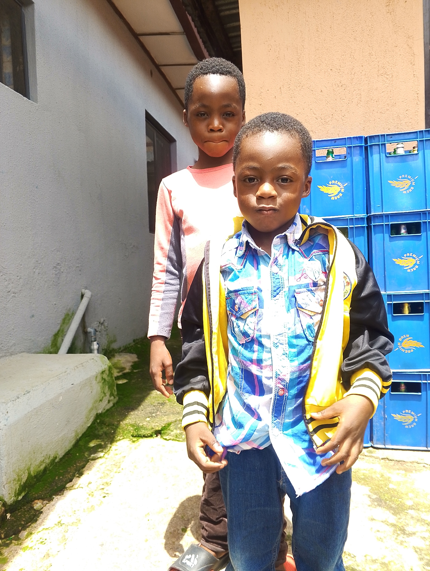 Two young boys stand outdoors. The boy in the foreground, on the right, wears a plaid shirt with a yellow and black jacket over it, and blue jeans. He is looking towards the camera. The boy behind him, on the left, wears a long-sleeved pink and grey shirt and dark pants. He is also looking towards the camera. They are standing on a concrete surface in front of a light-colored building with a dark roofline. To the right of the boys are stacked blue plastic crates, some with yellow logos on them. A concrete planter box with green moss growing on it is visible to the left. The scene appears to be in an urban setting.