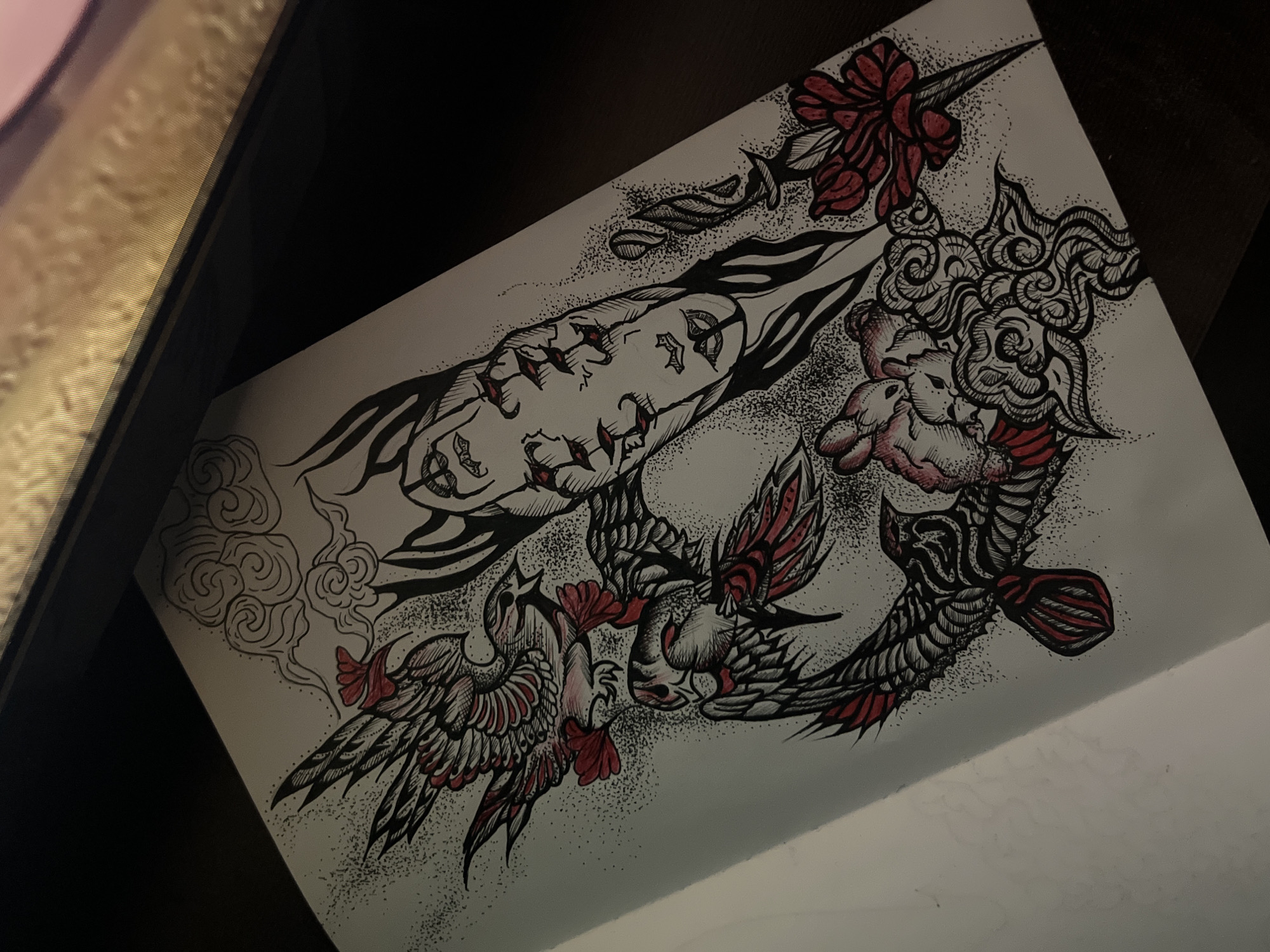 An intricately detailed, stylized drawing is depicted on a sheet of white paper, captured from an angled, slightly elevated perspective. The artwork is executed primarily in black ink with striking red and subtle pink accents.
The central subject on the upper left is an elongated, distorted human-like face, characterized by exaggerated features, flowing dark tendrils or hair, and prominent stitch-like marks along the cheek and jawline. Extensive stippling creates dense shading and a textured backdrop around this figure.
Adjacent to the face, towards the upper right, are intricate, swirling cloud patterns, strongly reminiscent of traditional East Asian (irezumi) art. Intertwined with these clouds and other elements are two small, cherubic figures with soft pink shading, along with vibrant red floral or ribbon-like designs and sharp, pointed motifs.
In the lower and central sections of the composition, two fantastical bird-like creatures are prominently featured. Their wings are elaborately drawn with bold black outlines, dark shading, and red and pink accents, conveying a sense of dynamic movement. More of the traditional cloud patterns and widespread stippling surround these birds, contributing to an atmospheric, almost smoky effect.
The artwork blends elements of dark art, traditional Japanese irezumi cloud motifs, and a neo-traditional aesthetic. The limited yet impactful color palette emphasizes the black linework and stippling against the vibrant red and subtle pink details. The paper rests on a dark, flat surface, likely a desk, with a blurred, lighter object in the upper left background, indicating an indoor setting. There is no visible text in the image. The overall impression is dramatic and somewhat unsettling, stemming from the stylized distortion of the face and the intense interplay of the various design elements.