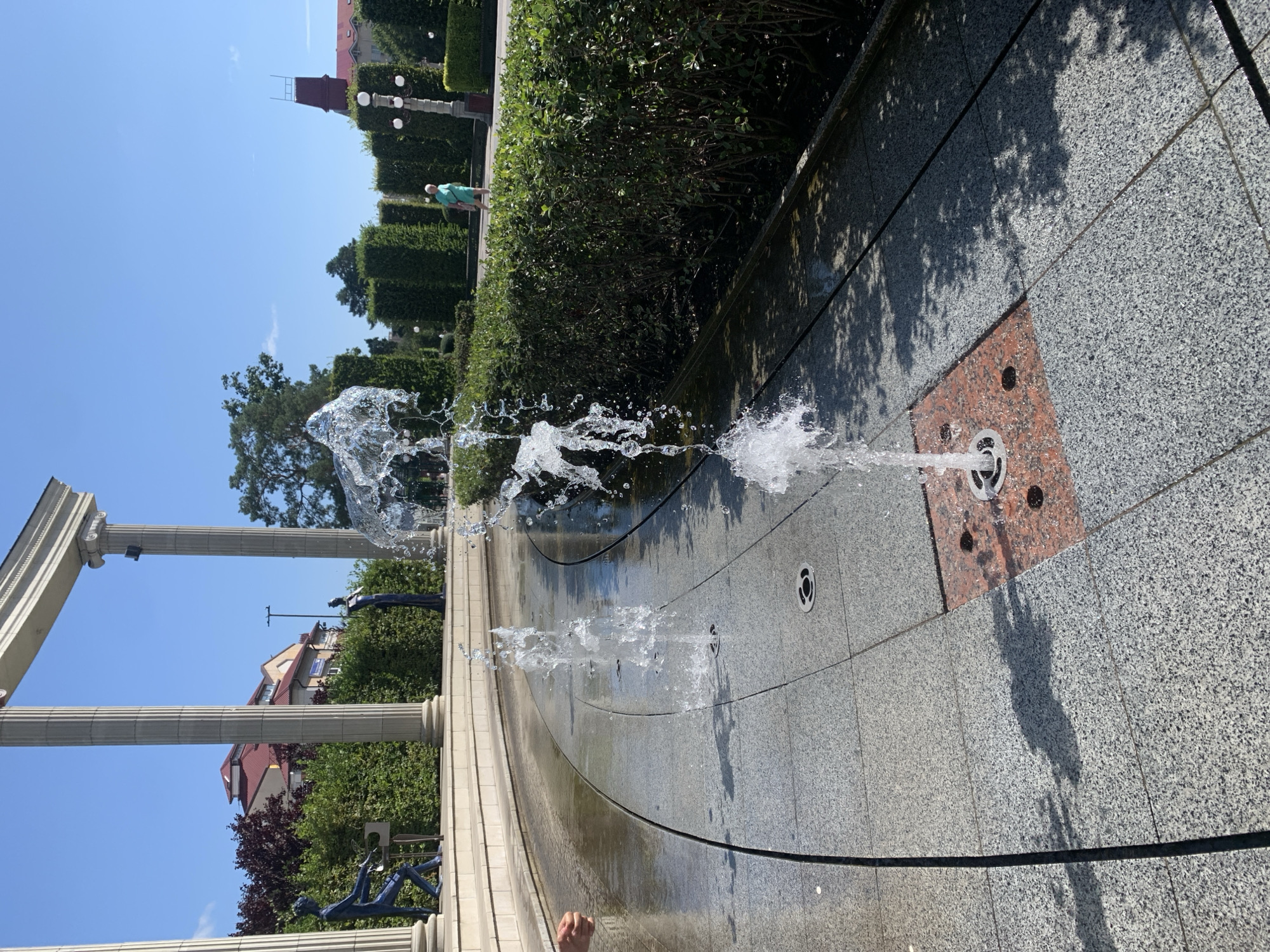 The image depicts a public space in Irpin, Ukraine, featuring a decorative fountain with water jets spraying upwards. The fountain is situated on a patterned paved area with granite tiles and a reddish insert around one of the water outlets. To the left, a curved, wet path reflects the sky and surrounding elements. In the background, manicured green hedges form a backdrop for a walkway where a person in a turquoise outfit is walking away from the camera. Beyond the hedges, trees and buildings are visible under a clear blue sky. Several large, classical-style columns stand to the left, framing a view of buildings with red roofs. Two statues, one of a figure seated at a piano and another standing, are positioned behind the hedges and columns. The overall scene suggests a well-maintained park or plaza designed for public enjoyment.