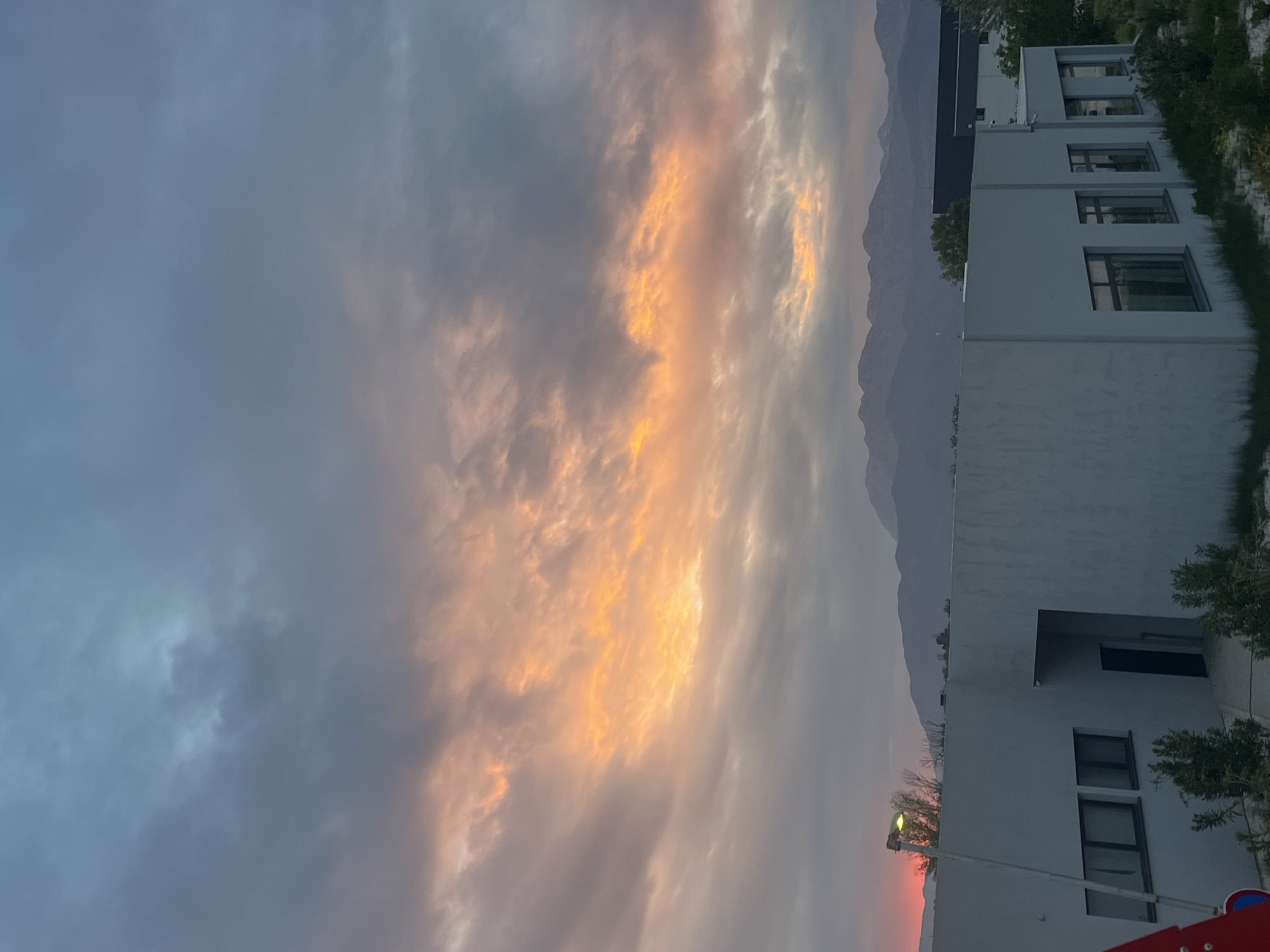 The image shows a cityscape at dusk in an unknown location. Mountains form a backdrop to modern white buildings on the right side of the frame. The sky is predominantly filled with clouds illuminated by the setting sun, casting an orange and yellow glow. A streetlight is visible in the lower right, along with parts of playground equipment. The overall scene is calm and serene.