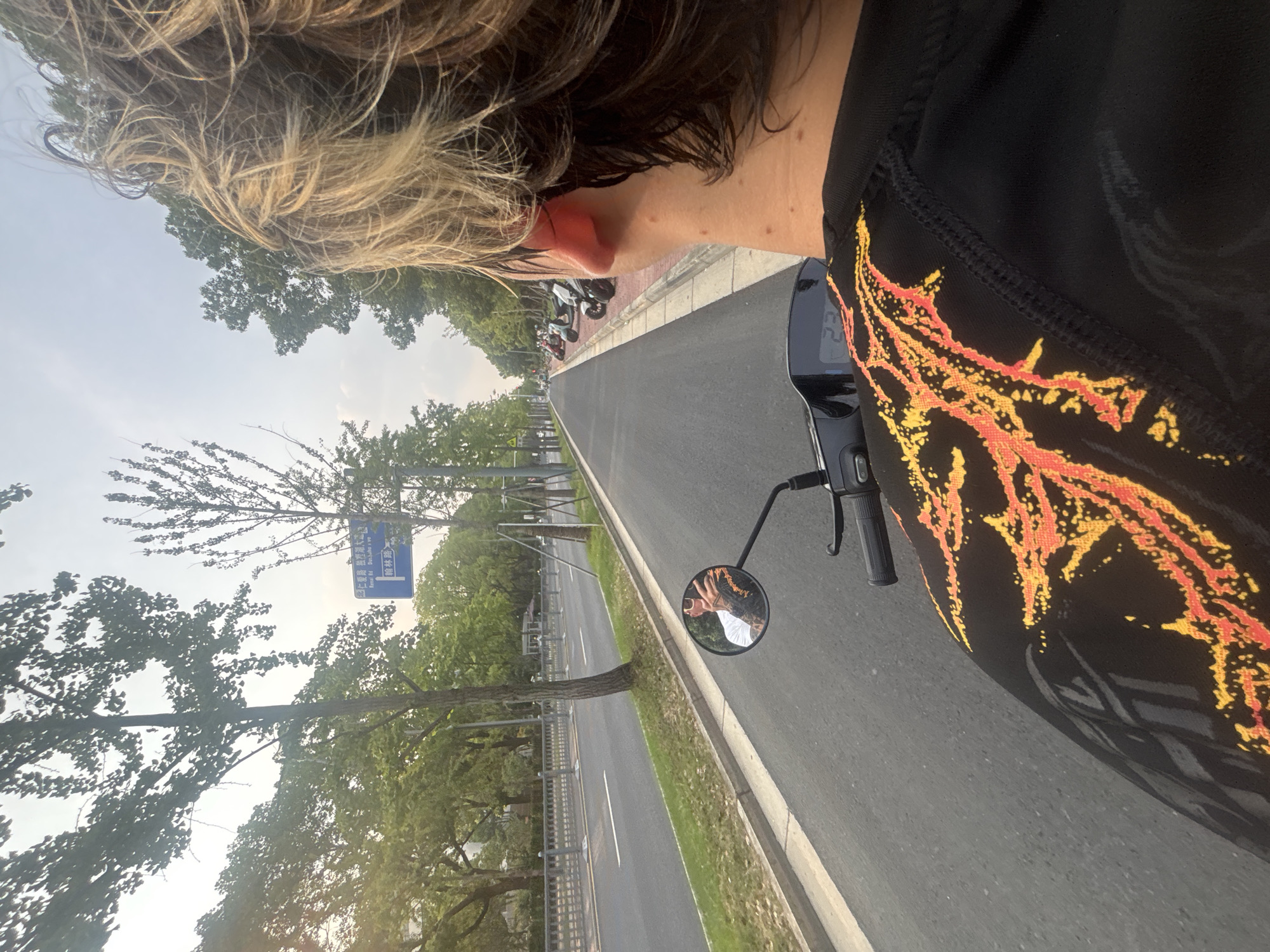 The image shows a first-person perspective of someone riding a scooter on a paved road in Suzhou Industrial Park, China. The rider's head and shoulders are visible in the foreground, with their blonde hair blowing in the wind. They are wearing a black shirt with an orange and yellow flame-like pattern. A mirror on the scooter's handlebars reflects the rider's hand and arm. The road is lined with trees and a sidewalk. In the distance, more scooters are parked along the side of the road. A blue street sign with Chinese characters is visible on the left side of the road. The sky is overcast.