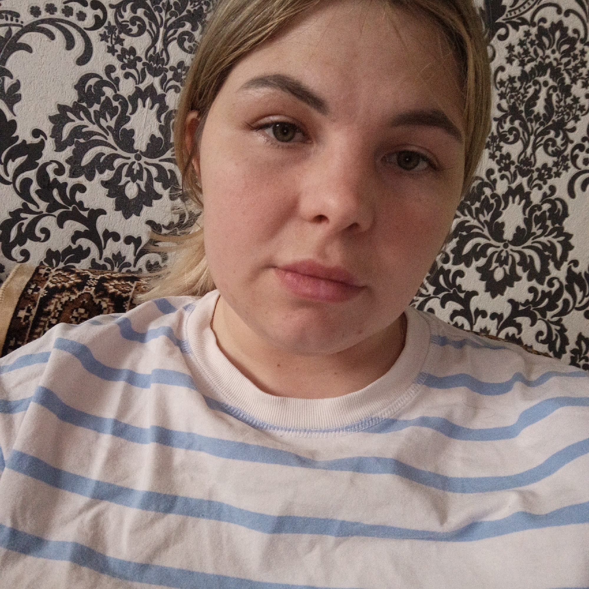 A young woman is captured in a close-up, chest-up shot, looking directly at the camera. She has light blonde or light brown hair pulled back, fair skin, and green/hazel eyes. Her eyebrows appear well-defined. Her facial expression is neutral, calm, and slightly pensive.
She is wearing a simple, white t-shirt with evenly spaced light blue horizontal stripes and a ribbed white collar. The fabric of the t-shirt appears soft and slightly rumpled.
The background consists primarily of a wall covered in light-colored wallpaper featuring prominent, intricate dark (likely black or dark grey) floral and damask-like patterns. The wallpaper has a slightly textured appearance. To the viewer's right, behind her left shoulder, a portion of a patterned fabric item, possibly a cushion or blanket, is visible. This fabric has a more rustic or tribal pattern with shades of brown, black, and beige.
The scene takes place indoors, likely within a home, suggested by the wallpaper and soft furnishing. The lighting is consistent, suggesting a well-lit interior, but the time of day is not discernible. No specific location cues are present beyond the domestic setting. There is no visible text in the image.