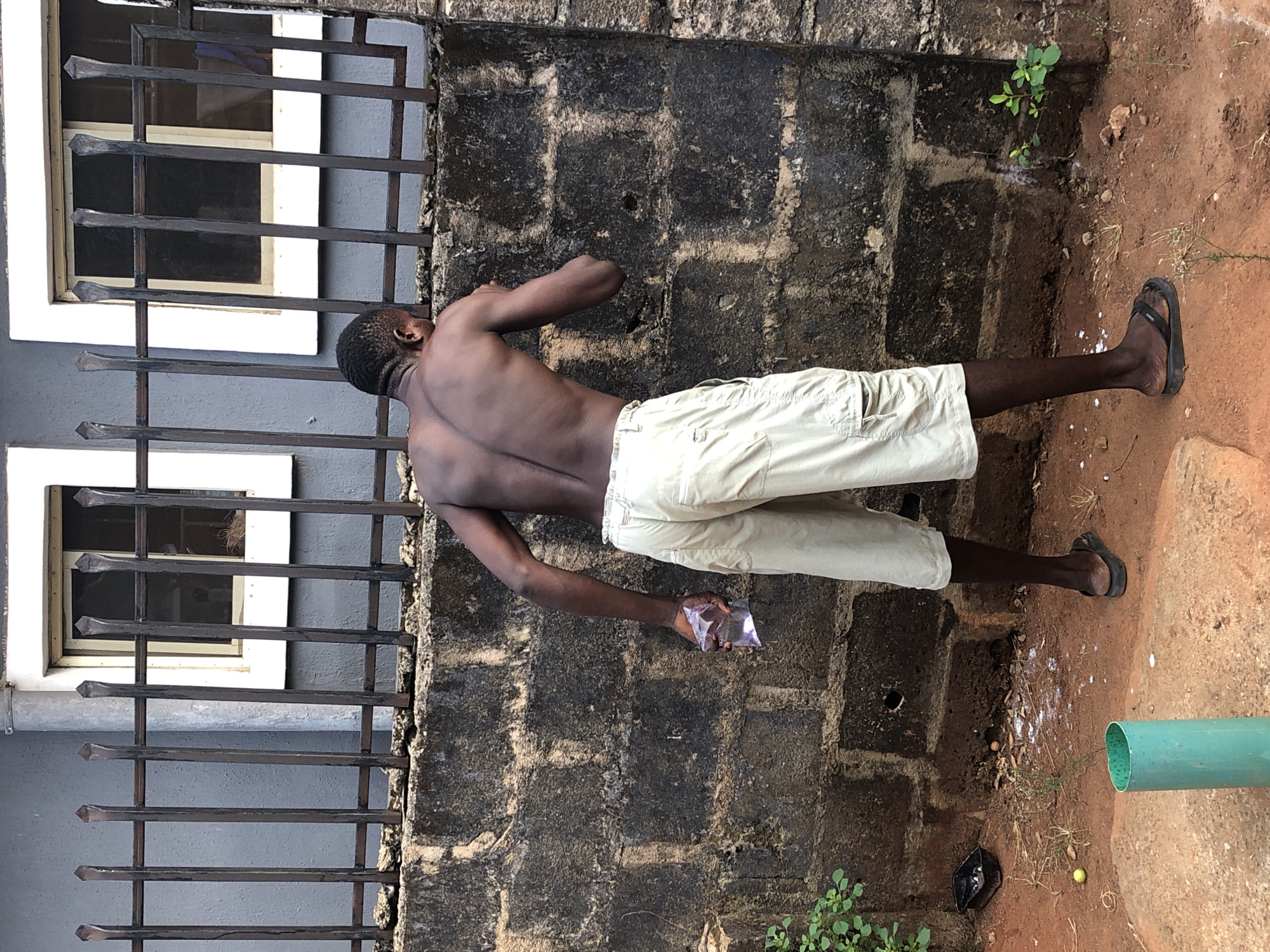 A person stands adjacent to a brick wall and a window with iron bars, holding a plastic bag. The scene includes elements like plants, sandals, and a green pipe on the ground. The location is not specified beyond "Unknown, Unknown," so it's not possible to pinpoint the city.