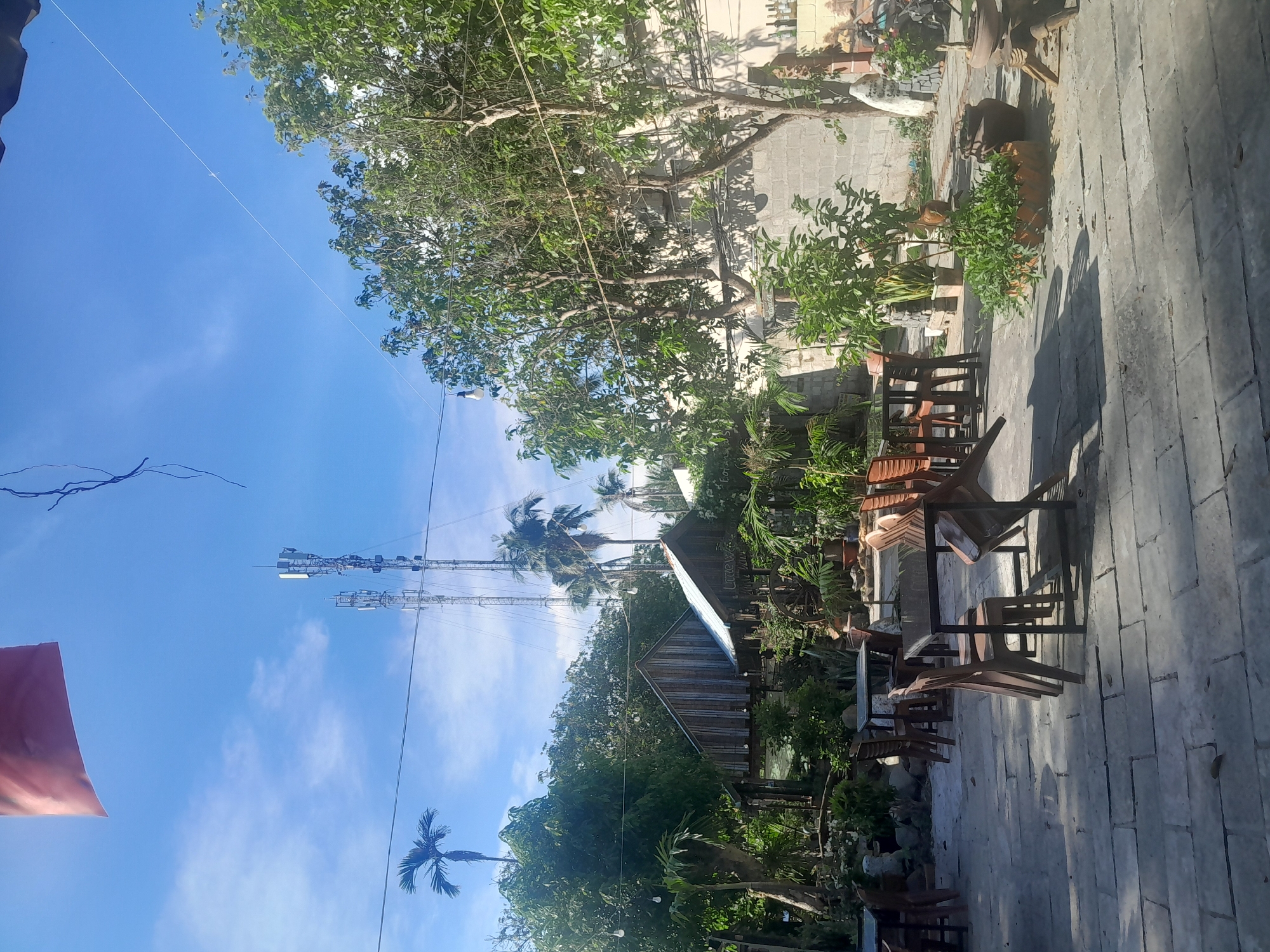 The image depicts an outdoor seating area of a restaurant, likely in the city of Phan Thiet Ward, Vietnam, given the presence of palm trees and a tropical-style garden. The area is paved with large, light-colored stone tiles, with tables and chairs set up for dining. Several tables are visible, some made of dark wood with metal legs and others appearing to be glass-topped. The chairs are mostly brown plastic, with a few in a lighter shade.
The seating area is surrounded by lush greenery, including various trees, potted plants, and flowering bushes. Two wooden structures, resembling small huts or shelters, are present in the background. One structure has a weathered wooden facade and a corrugated metal roof, while the other, situated to its right, also has a wooden exterior and a metal roof, with the word "Little" visible on a sign attached to it.
In the distance, two tall cellular towers stand against a clear blue sky with scattered white clouds. A red flag or banner is partially visible in the upper left corner of the image. Overhead, strings of lights are strung between trees, suggesting the area is used in the evening. The overall ambiance is relaxed and natural, indicative of a casual dining establishment. No people are visible in the image.