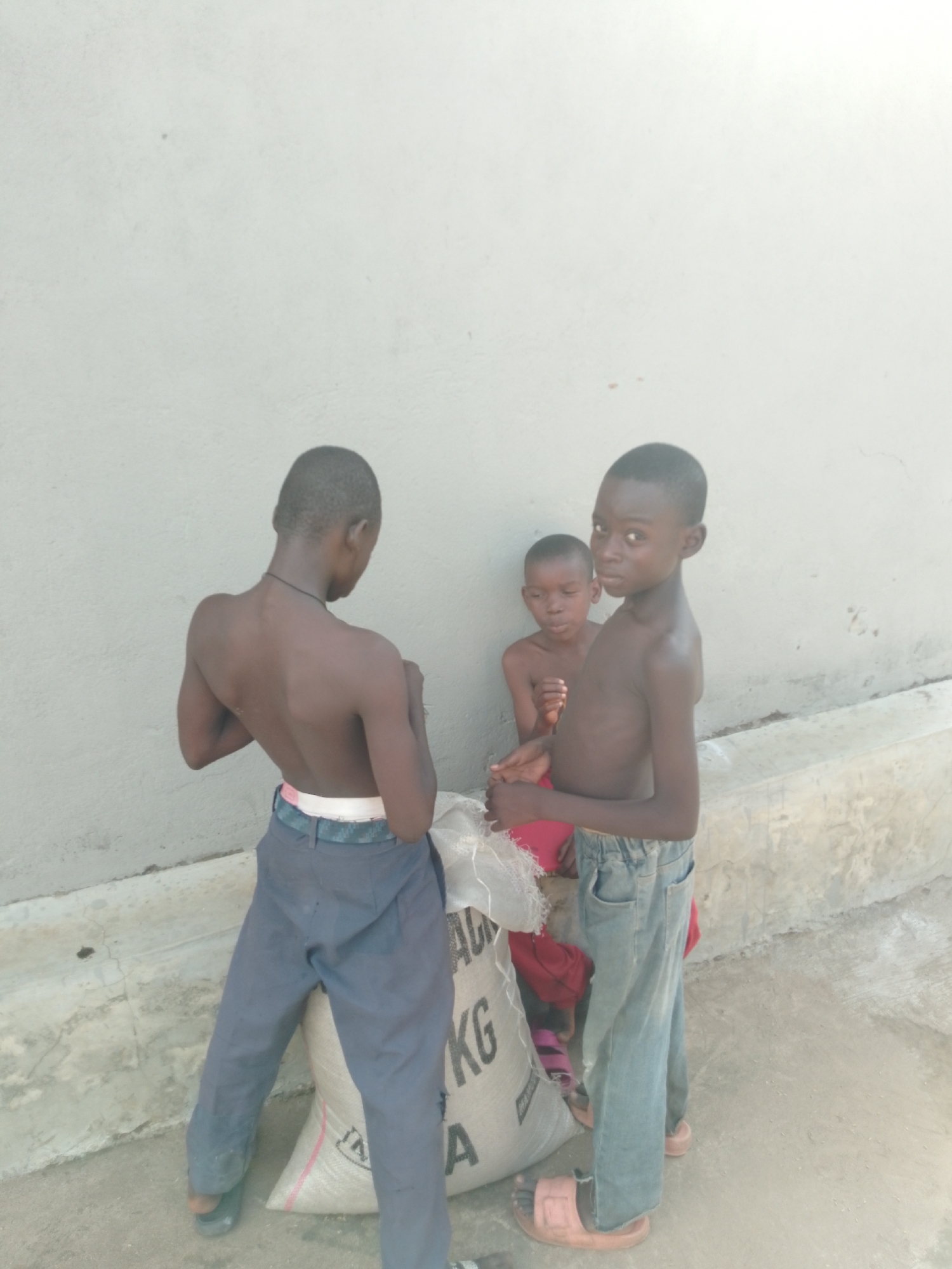 Three young boys are standing next to a large burlap sack against a light grey wall. The boy on the left, with his back to the camera, is wearing dark blue pants and a white belt. He has his arms bent and is looking over his left shoulder. The boy in the middle is wearing a red shirt and is leaning against the sack. The boy on the right is facing the camera, wearing light blue jeans and pink sandals. He is looking directly at the camera with a serious expression. The burlap sack appears to be filled with grain and has lettering on it, though it is partially obscured by the boys. The ground is concrete. The scene appears to be in an outdoor area, possibly a courtyard or alleyway, in Serti, Nigeria.