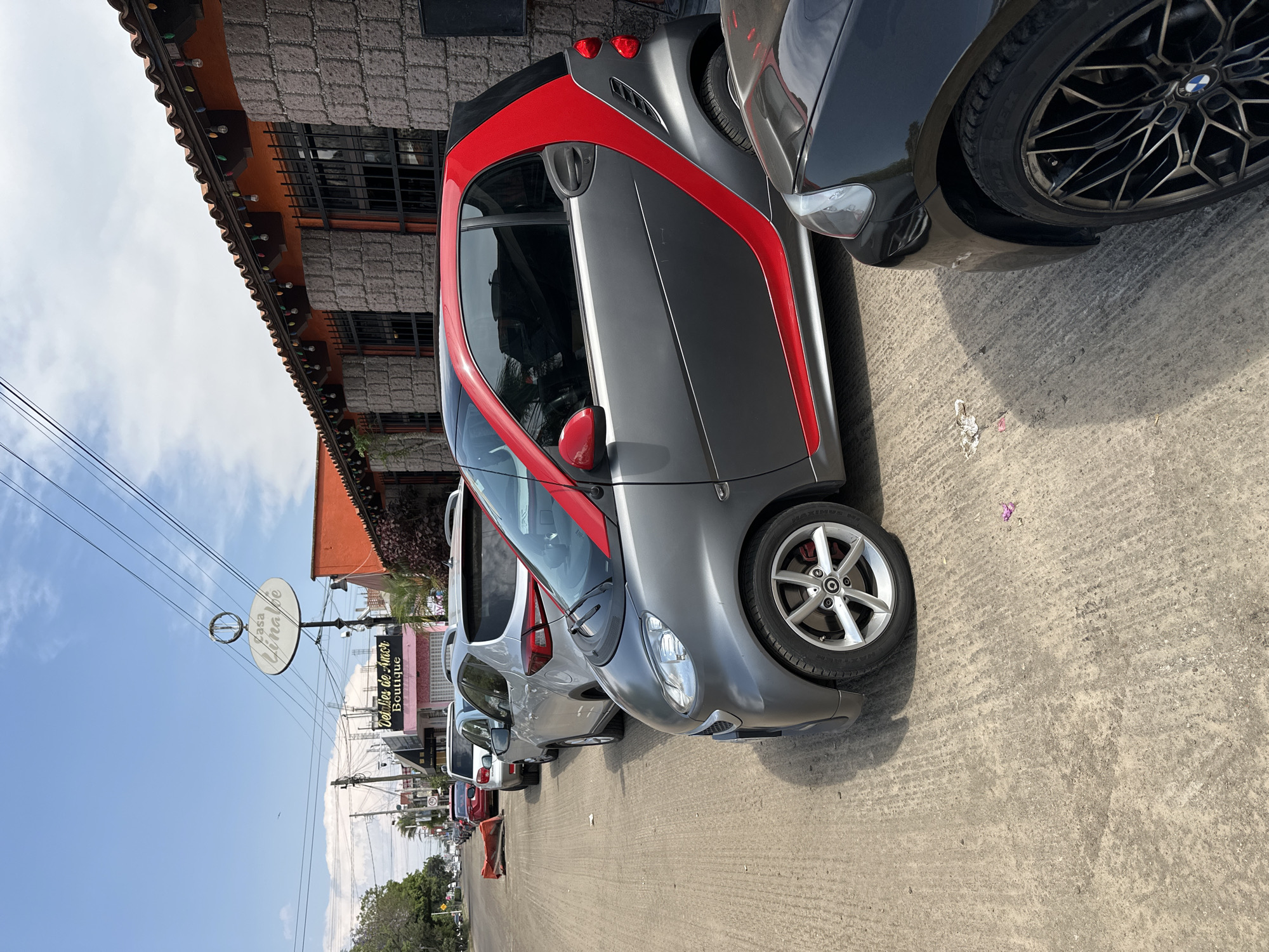 The image shows a row of parked cars on a street. A gray and red Smart car is the most prominent vehicle. The scene is in Puebla, Mexico, indicated by a sign reading "Casa Yinape" and "Detalles de Amor Boutique". A woman is standing further down the street. The sky is blue with some clouds, and power lines are visible overhead. The buildings have a tiled and concrete exterior.