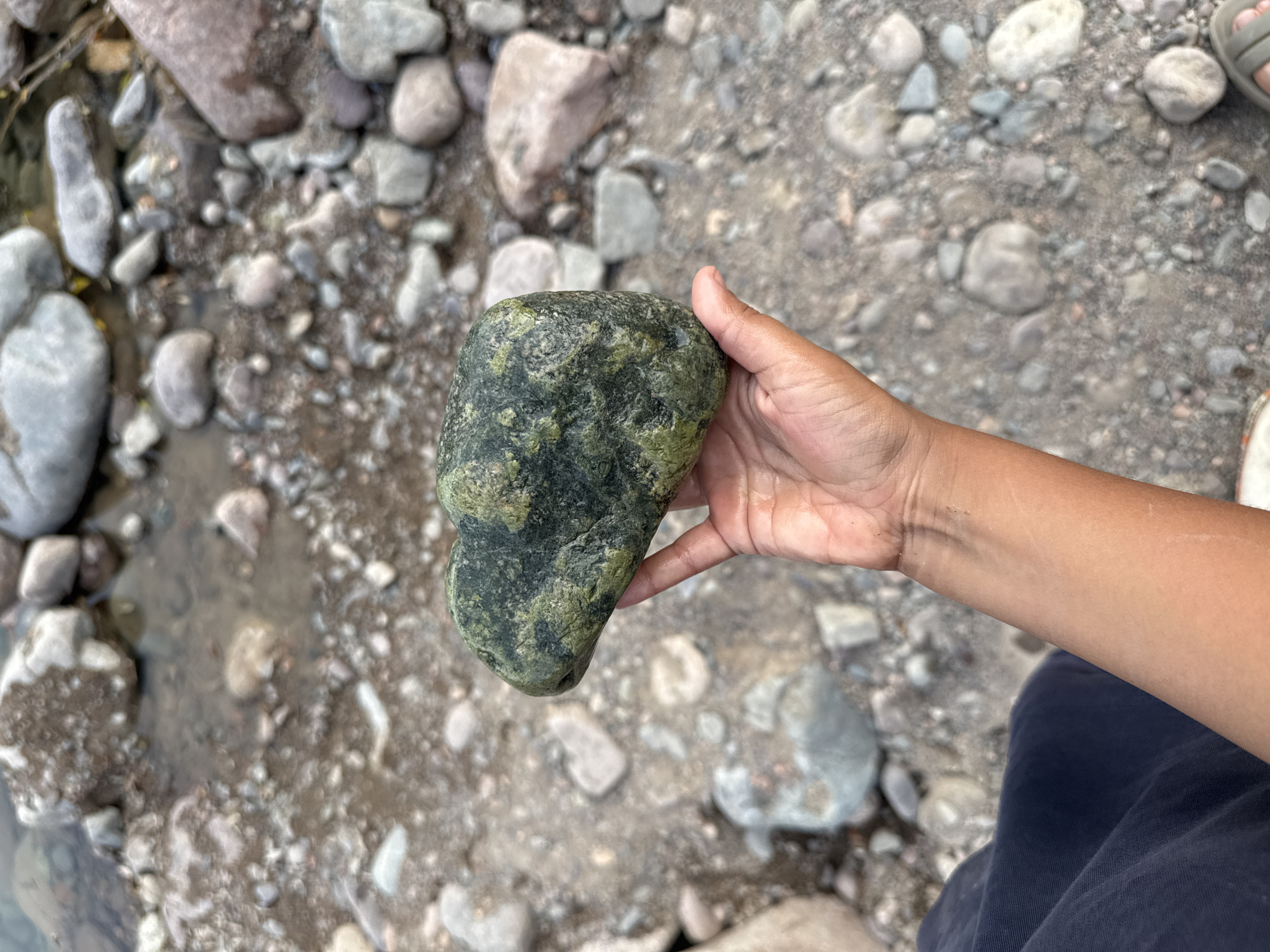 A close-up, eye-level shot captures a hand holding a dark green, irregularly shaped rock. The rock has a rough texture with lighter green and black speckles, and it is wet. The hand is tanned with short fingernails and is positioned in the lower right of the frame. The background is a blur of river rocks and shallow water, suggesting a natural outdoor setting. The lower left of the image shows a dark navy blue fabric, likely clothing. The scene appears to be in Leskhoz, Kazakhstan.