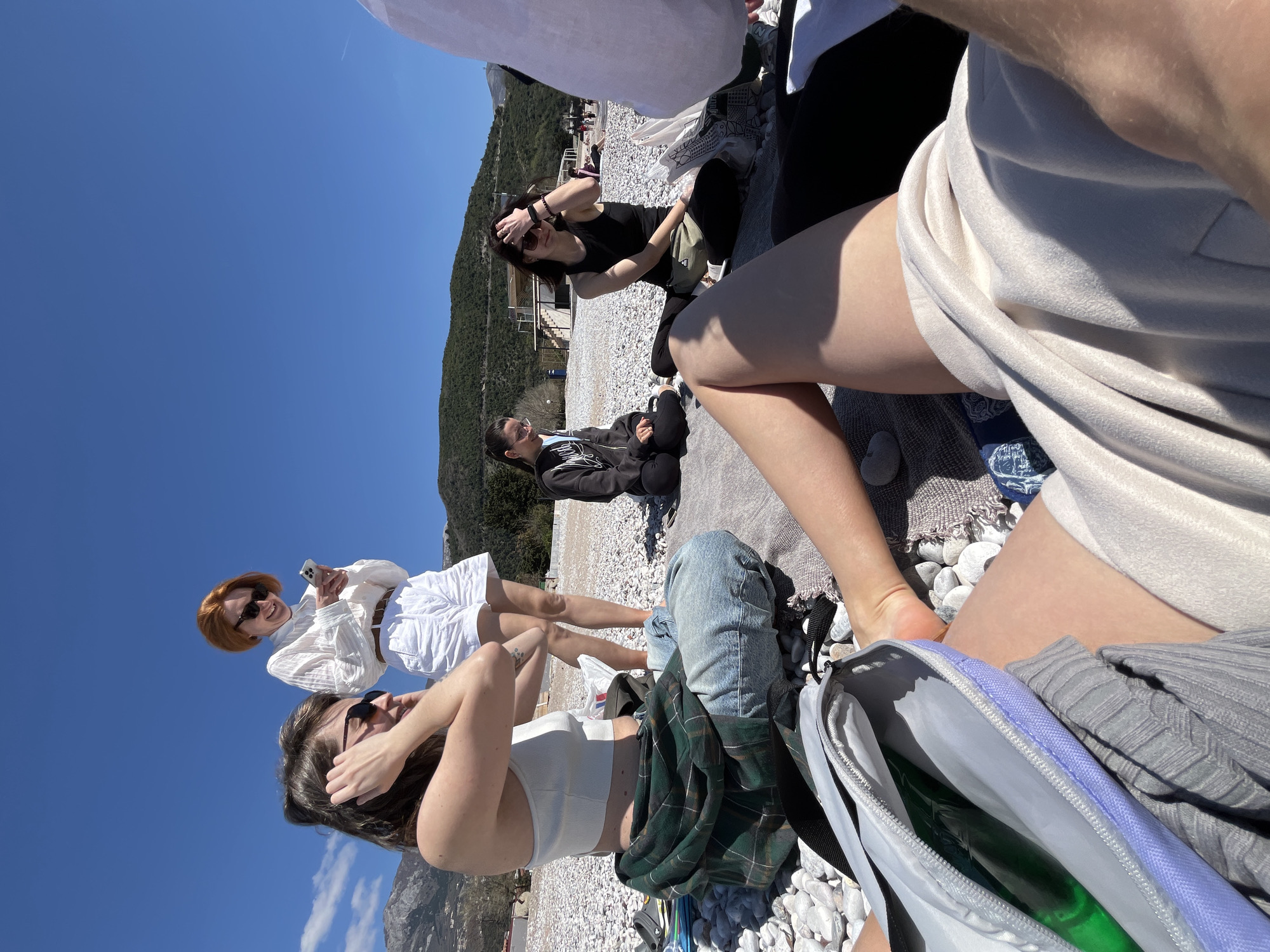 The image captures a group of five young women on a pebble beach. In the foreground, a woman in a white crop top, denim jeans, and a green plaid shirt sits with her hands touching her hair, while next to her, another woman with short red hair, sunglasses, and a white outfit holds a smartphone. Behind them, a woman in a black top has her hand on her forehead. Further back, a fourth woman in a black hoodie is sitting on a blanket. In the background, a tree-covered hillside rises, and the clear blue sky is visible above. To the right of the image, a person sits with their legs and gray shorts visible. To the right is a light gray bag. The location is likely a beach near Budva, Montenegro, given the pebble beach, mountainous backdrop, and clear sky, which are characteristics of the Adriatic coastline in that region.