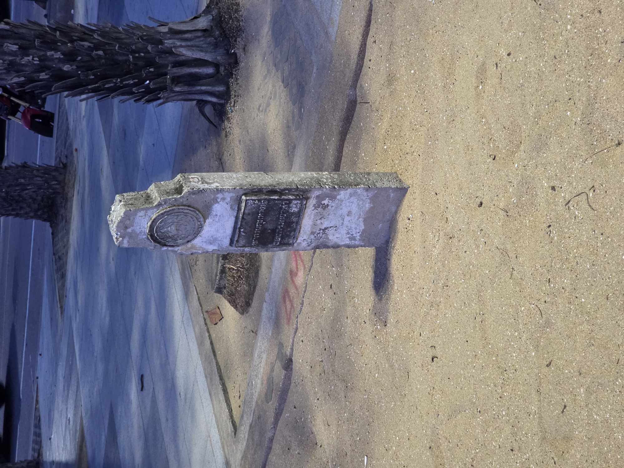 The image presents an outdoor scene in Pattaya City, Thailand, under bright daylight conditions, likely late morning or afternoon given the yellow tint of the light and shadows. The central focus is a weathered concrete marker or monument, positioned on the boundary between a sandy ground and a paved area.
This concrete marker features a dark metallic plaque, which displays text in Thai script, though it is unreadable at this resolution. To the left of the plaque, a circular metallic medallion is embedded, showcasing an embossed emblem or symbol. The marker itself exhibits considerable wear, with chipped edges, cracks, and general discoloration, suggesting exposure to the elements over time.
The ground immediately surrounding the marker is sandy, extending into the foreground and to the right, sprinkled with small stones and bits of debris. To the left and in the background, several tall, rough-textured palm tree trunks are visible, confirming a tropical setting. Behind one of the palm trees, a red motorcycle is partially seen, hinting at nearby activity. The paved area, which could be a sidewalk or road, is a light blue-grey color and displays distinct geometric patterns. A concrete curb separates the paved surface from the sandy ground, and some red graffiti or markings are visible on this curb. The scene is static, depicting no specific ongoing activity.