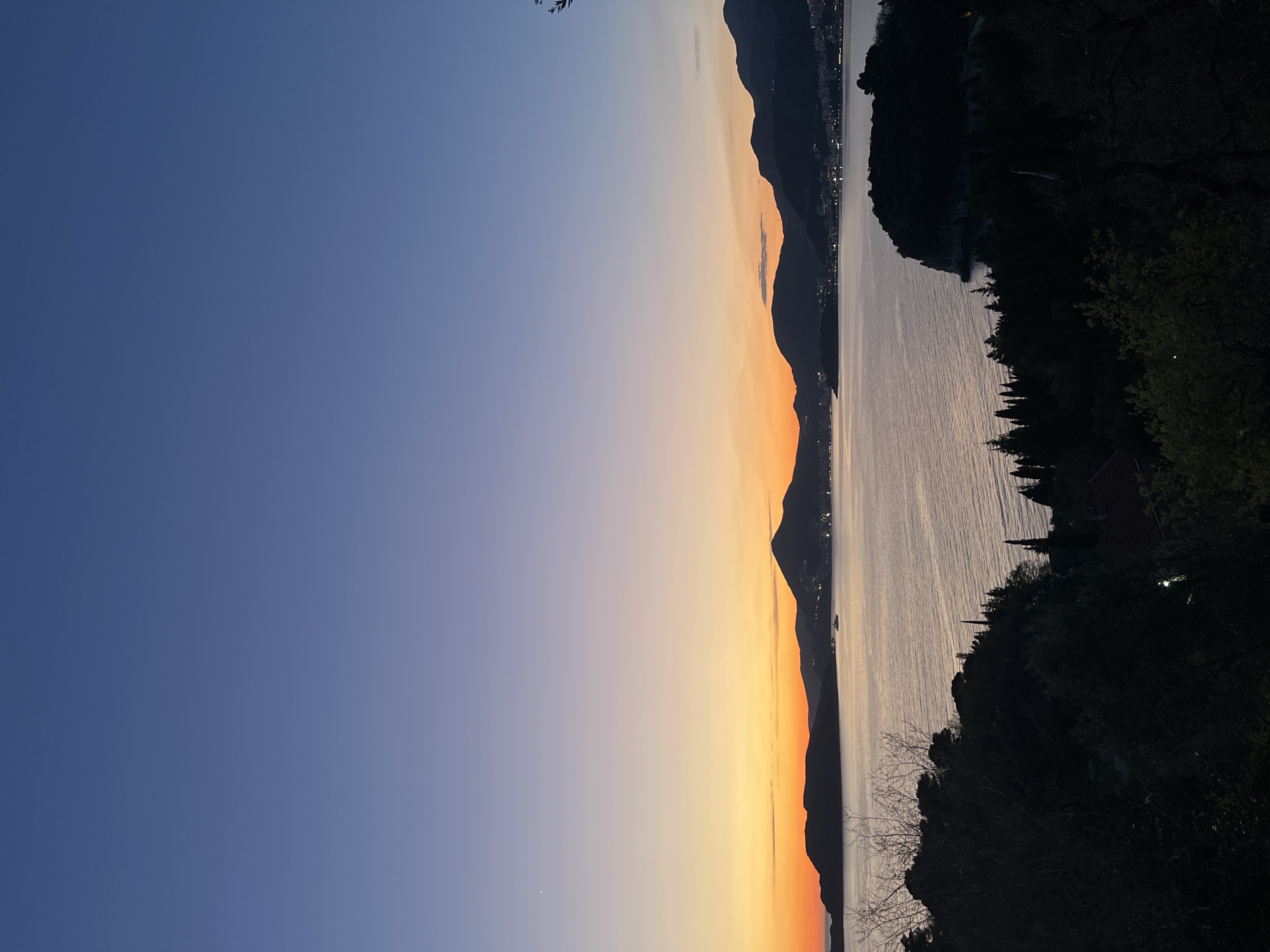 The image captures a twilight view in Sveti Stefan, Montenegro. The sky transitions from deep blue to orange over a mountain range silhouetted against the sunset. A body of water, possibly the Adriatic Sea, reflects the fading light. The coastline is partially obscured by dark foliage on the right side of the frame. A few lights are visible in the distance along the mountains.