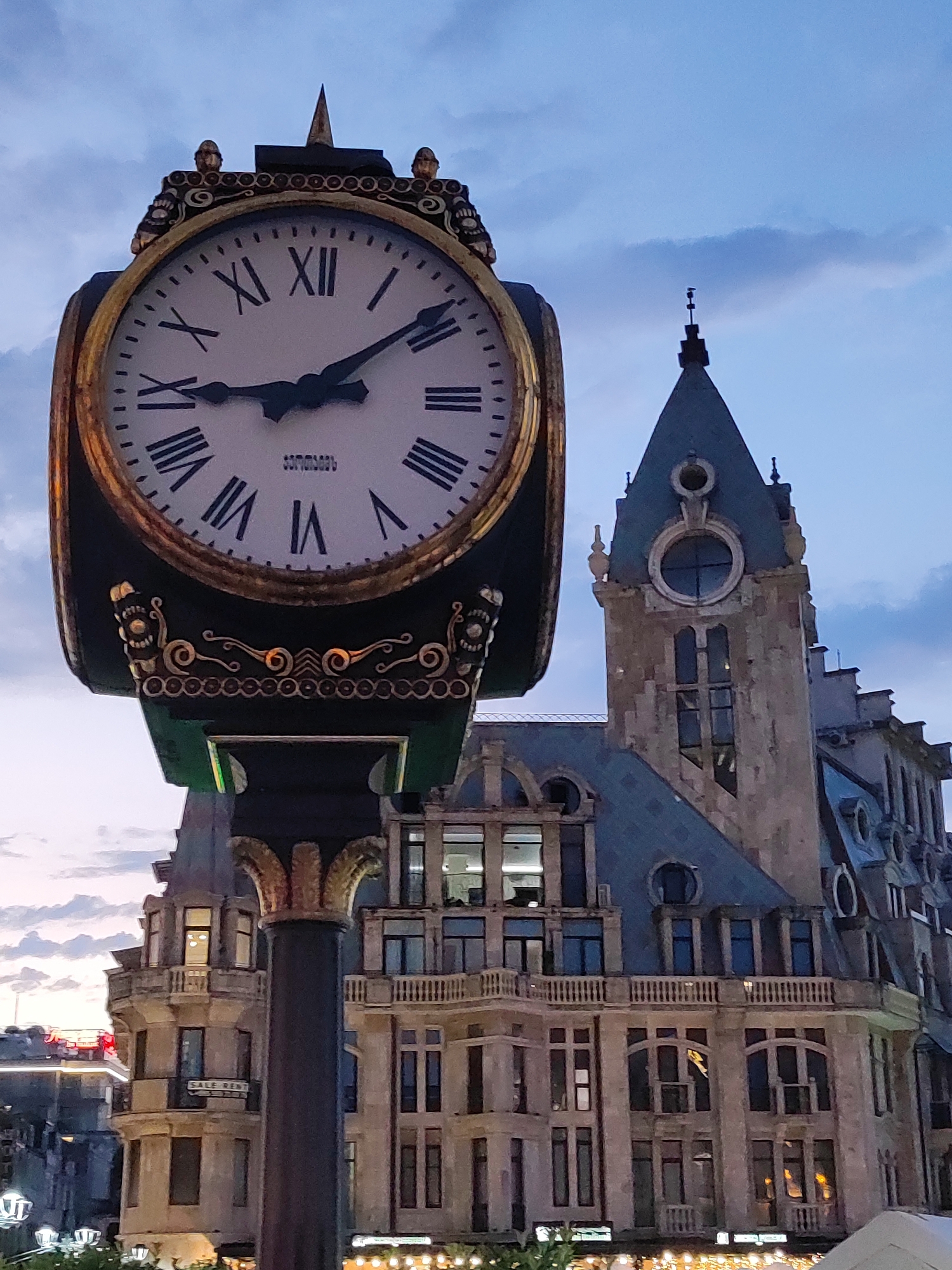 The image captures the iconic Batumi clock tower and a portion of the surrounding cityscape. The clock tower, a prominent landmark in Batumi, Georgia, stands tall with its ornate design and Roman numerals. The sky above is a dusky blue with scattered clouds, indicating it might be dusk or early dawn. The architectural style of the buildings suggests a European influence, typical of many structures in Batumi. The overall scene is one of urban beauty, with the clock tower serving as a central focal point against the backdrop of the city. There are no people or discernible actions visible in the frame.