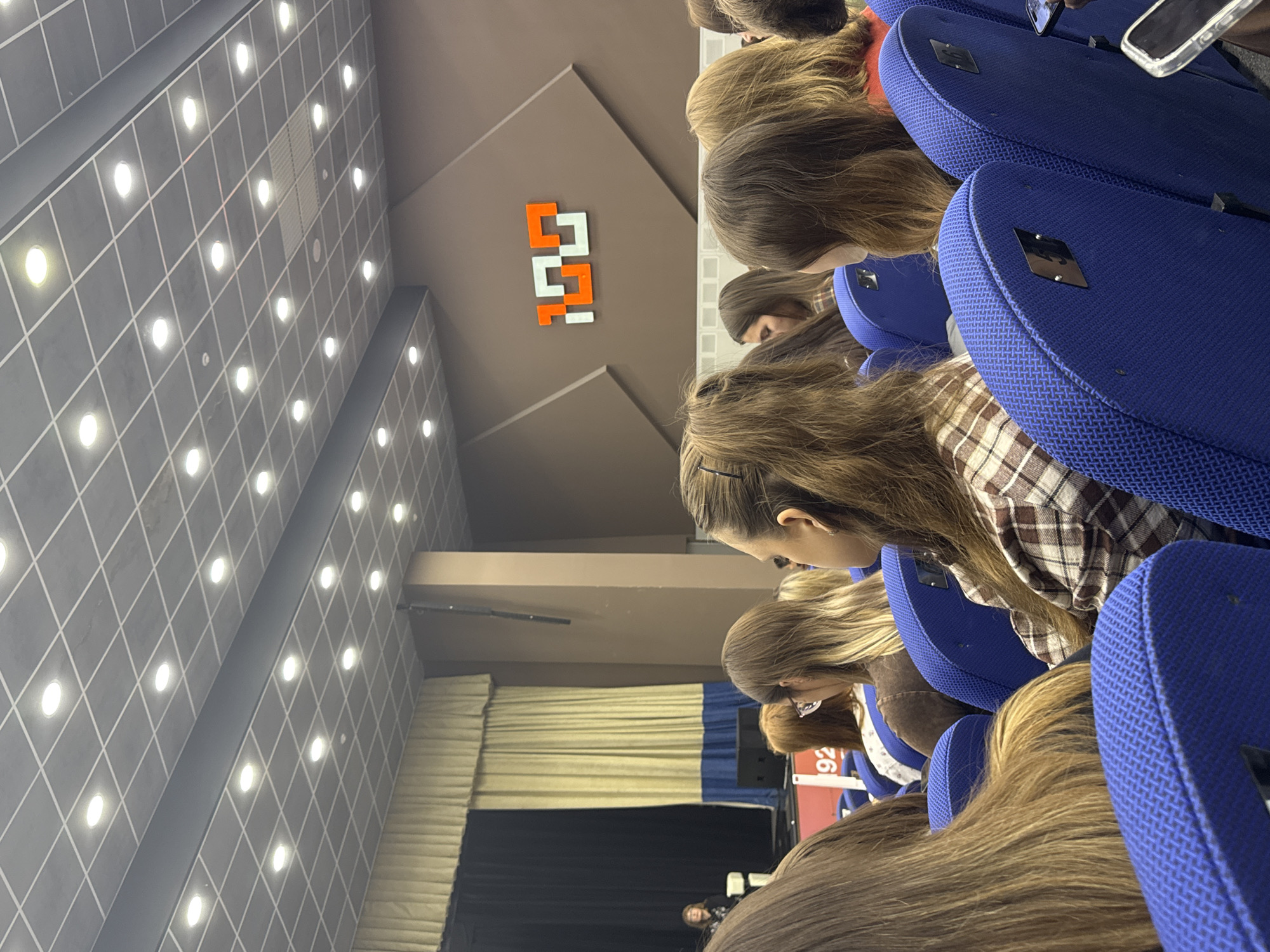 This image captures an interior view of an auditorium or lecture hall in Perm, Russia, filled with an audience. The perspective is from behind a row of attendees, looking towards the front of the hall and slightly upwards.
The primary subjects are numerous people, mostly young adults or students, seated in rows of chairs. Their backs and the tops of their heads, mostly with long, brown hair, are visible as they face forward, seemingly attentive to an event or presentation. Some individuals are seen in profile. In the distance, on a stage area, a single person is faintly visible, suggesting they are speaking or performing.
The setting is characterized by neat rows of blue, textured upholstered chairs. Mobile phones are visible in the seatback pockets or on the seats of some chairs. The ceiling features a grid pattern with evenly spaced, bright recessed lights. A large, geometric logo, composed of interlocking orange and white shapes, is prominently displayed on a brown wall to the right side of the main viewing area. The stage at the front is adorned with layered curtains, primarily a light yellowish-beige and a dark blue/black. On the back of one of the chairs, a white label with the number "92" is clearly visible.
The overall scene indicates an organized indoor event, such as a lecture, conference, or performance, taking place during what appears to be daytime or evening, given the reliance on artificial lighting. No specific emotions are discernible from the back views of the audience, but the arrangement suggests a focused atmosphere.