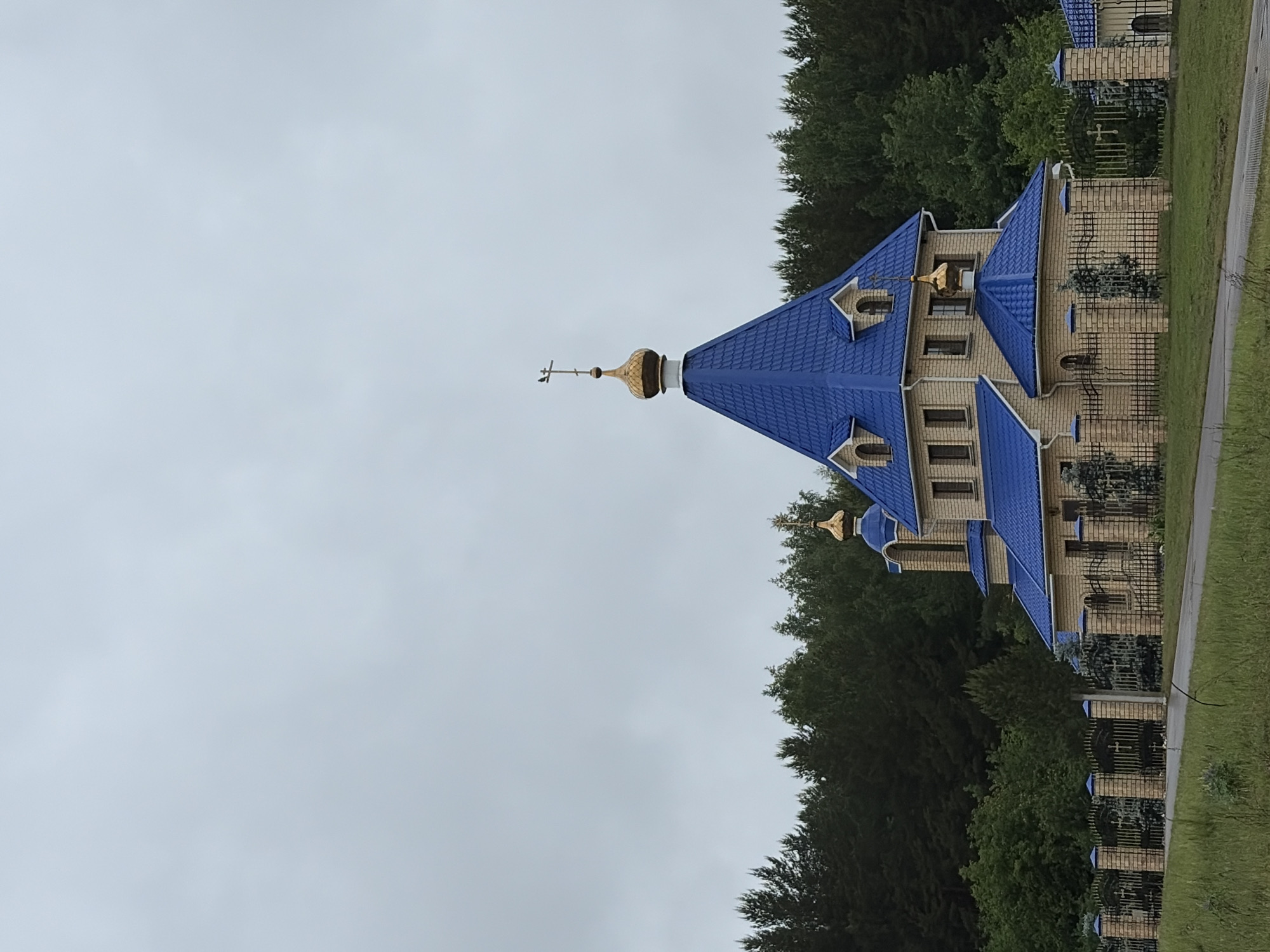 The image depicts a Russian Orthodox church located in Chaikovsky, Russia. The church has a blue tiled roof with multiple golden domes topped with Orthodox crosses. The building is constructed of light-colored brick with windows and is surrounded by a black metal fence. The church is situated next to a road and is bordered by a line of tall green trees. The sky is overcast.