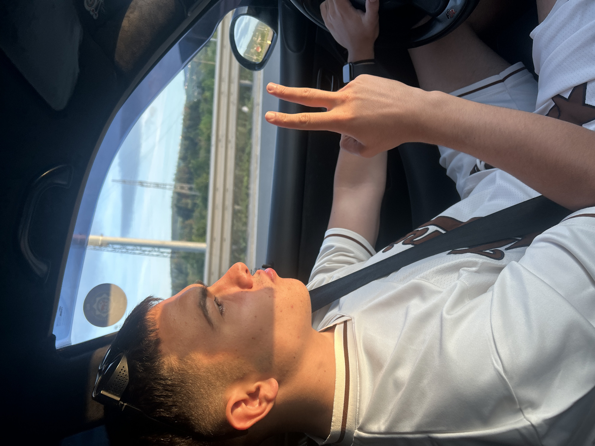 A young person is sitting in the passenger seat of a car, wearing a white jersey with brown trim and a seatbelt. They are turned towards the window, looking up, and making a peace sign with their right hand. Sunglasses are perched on their head. The driver's hand is visible holding the steering wheel. Outside the car window, a highway is visible with trees and a bridge in the background. A sticker is on the car window.