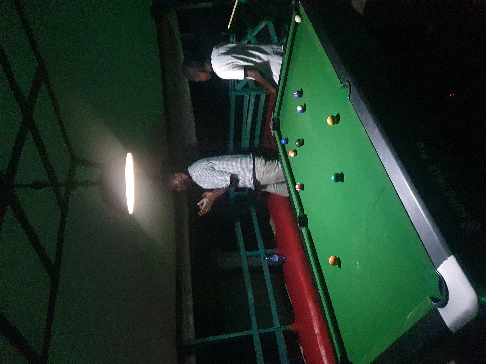 In Lokoja, Nigeria, a dimly lit pool hall is captured, focusing on a green pool table with several balls scattered across its surface. Two individuals are present. One person, wearing a checkered shirt and light-colored pants, stands to the left, appearing to gesture. To the right, a person in a white t-shirt is visible, holding a pool cue. The environment features a green-railed partition behind the table, and a red-upholstered bench. Overhead, a single lamp illuminates the scene. The pool table itself is branded "SUPERMAX Pro" along its side.