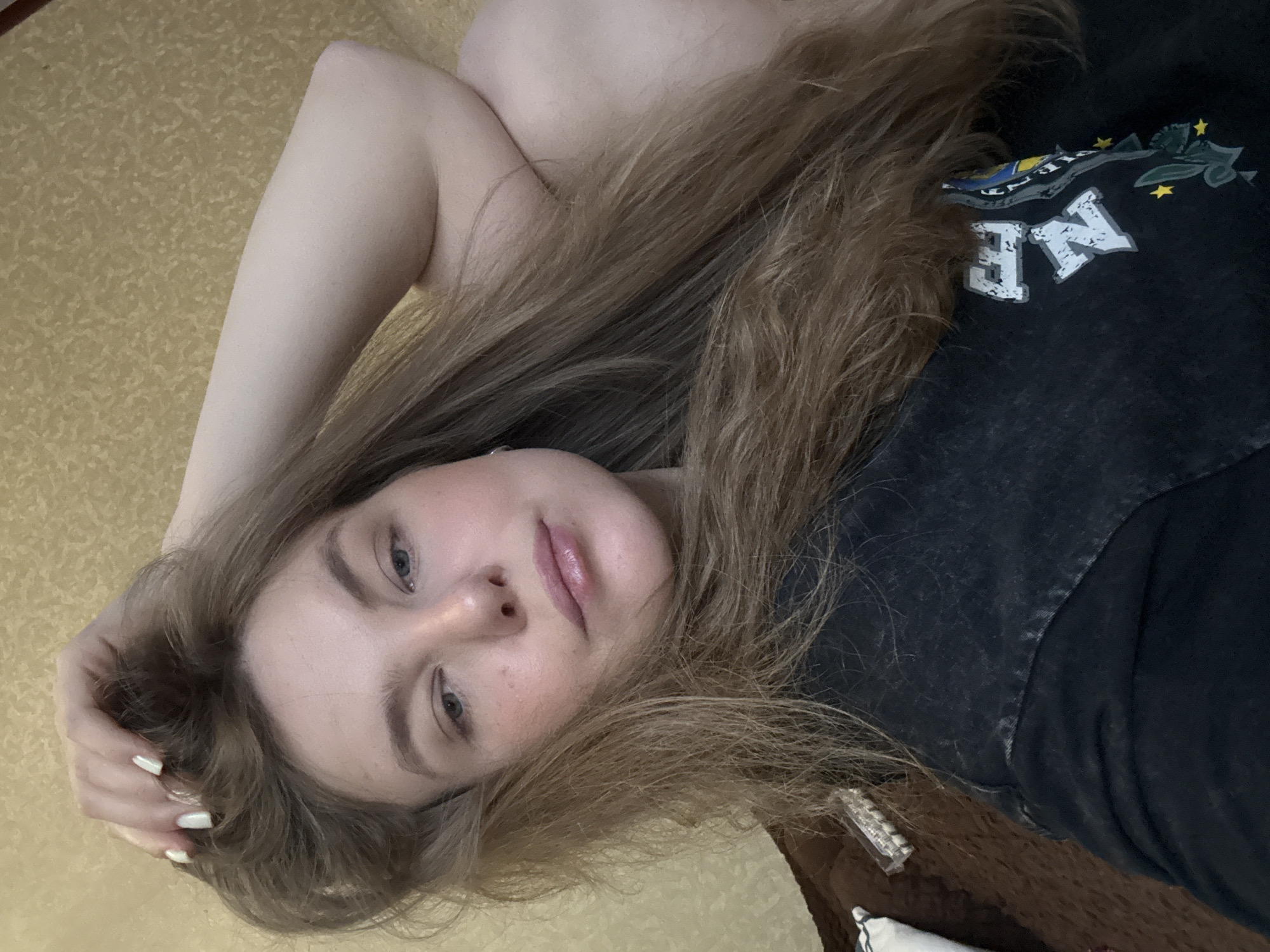 A woman with long blonde hair is lying on her back on a patterned surface, possibly a bed or floor. She is wearing a black t-shirt with some writing and design. Her left arm is raised above her head. The location is not discernable from the image.
